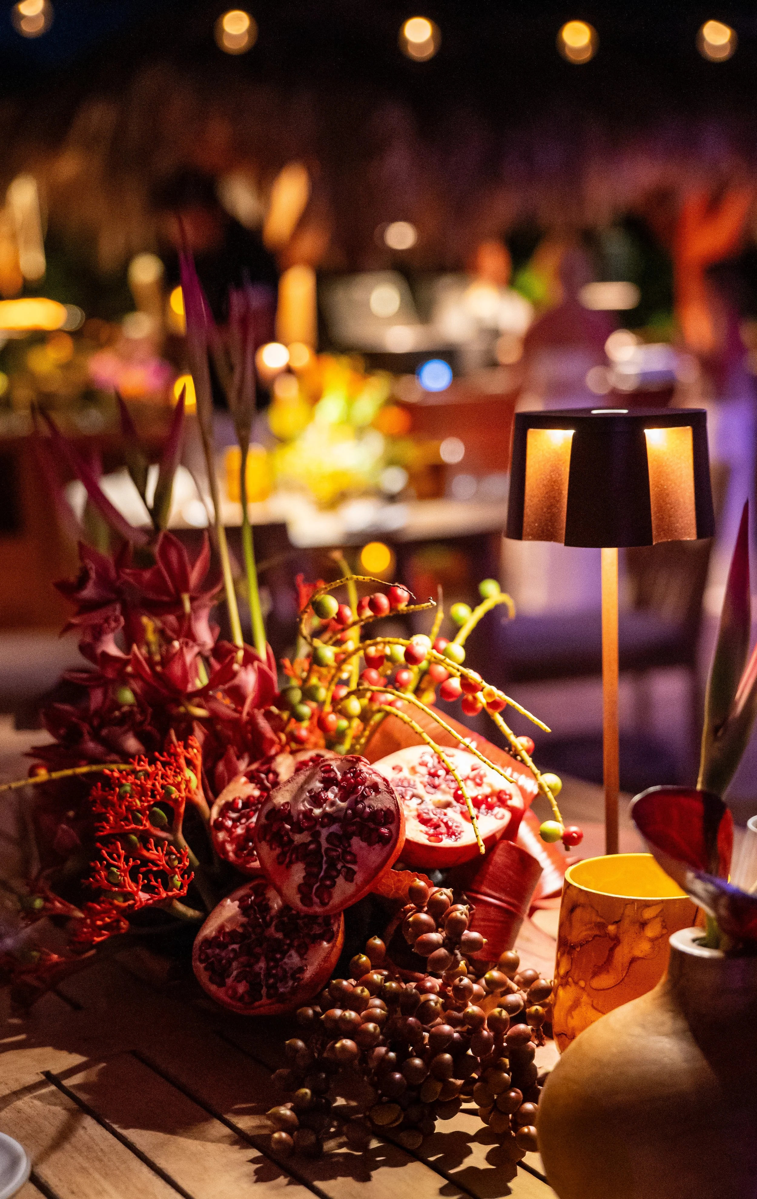 Décoration de table avec des fruits pomegranate, des branches de baies, et des vases. L'éclairage est tamisé avec des lumières chaudes en arrière-plan, créant une ambiance chaleureuse et festive.