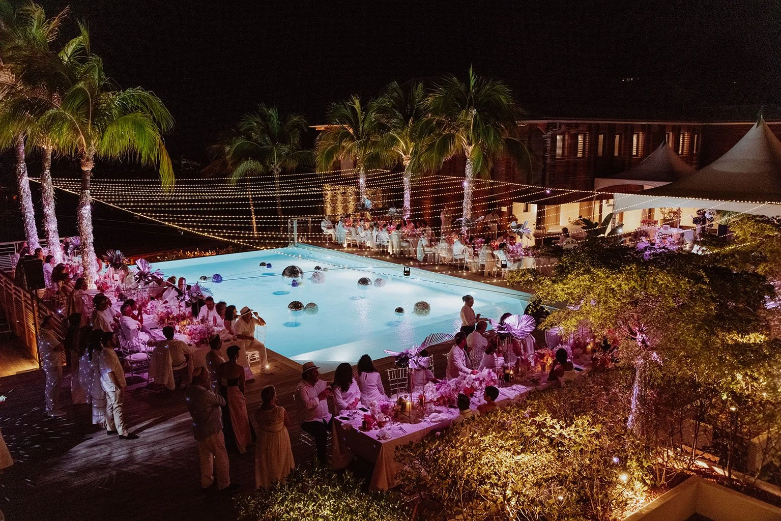 Une fête ou un dîner en plein air autour d'une piscine la nuit, avec des tables décorées de fleurs et de lumières, entourée de palmiers et de tentes.