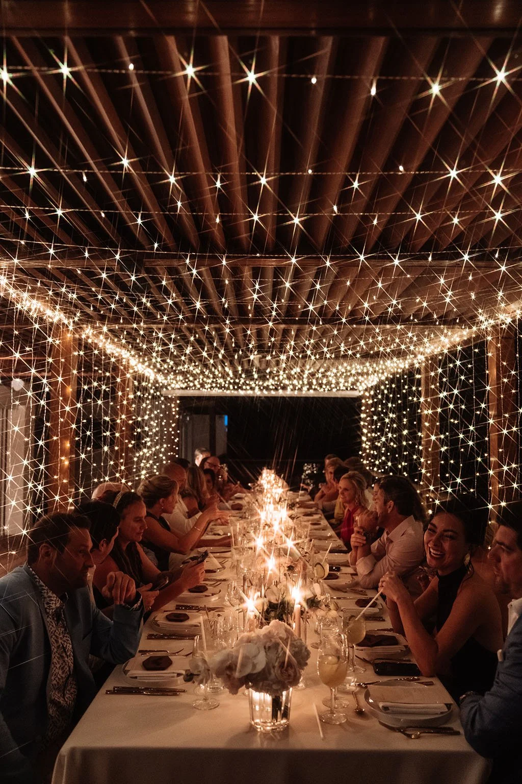 Groupe de personnes assises autour d'une longue table décorée, sous un plafond orné de guirlandes de lumières scintillantes dans un cadre chaleureux et festif.