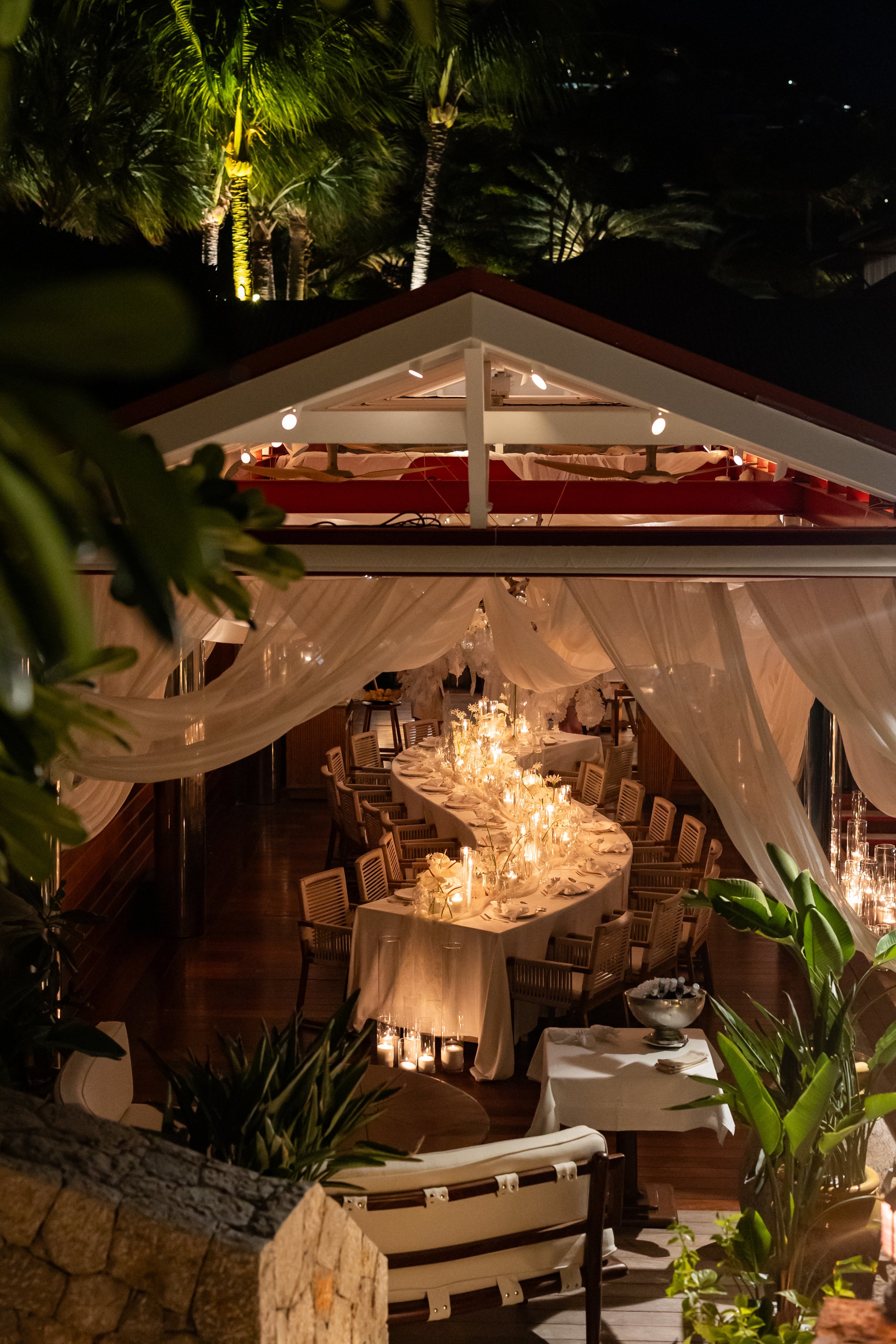 Une salle à manger décorée pour un dîner, avec une longue table drapée d'une nappe blanche, entourée de chaises en bois, éclairée de bougies et de chandeliers.