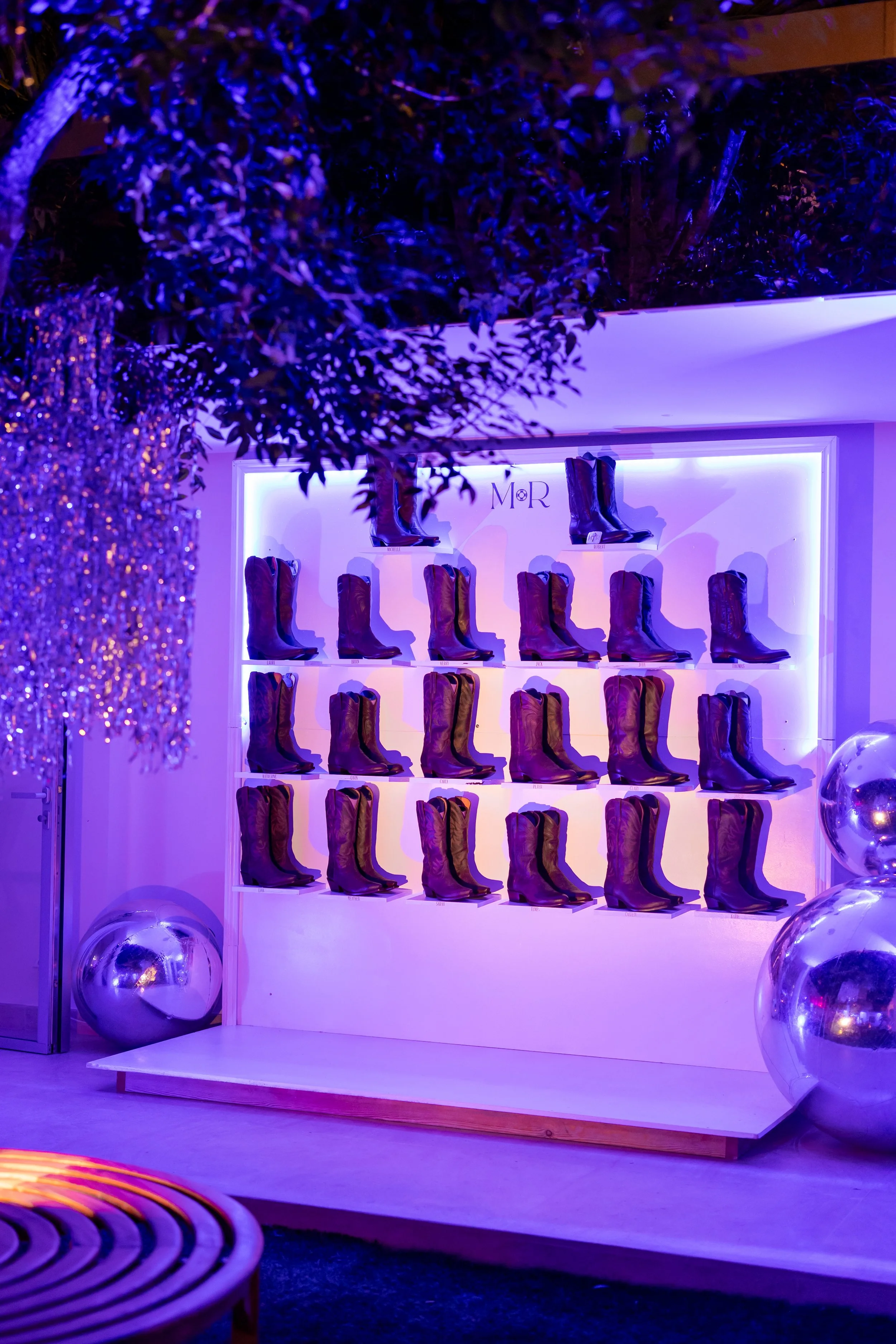 Vitrine avec des bottes en cuir, éclairée par un éclairage violet, décorée avec des ballons argentés et des éléments de décoration en apportant une ambiance élégante et moderne.