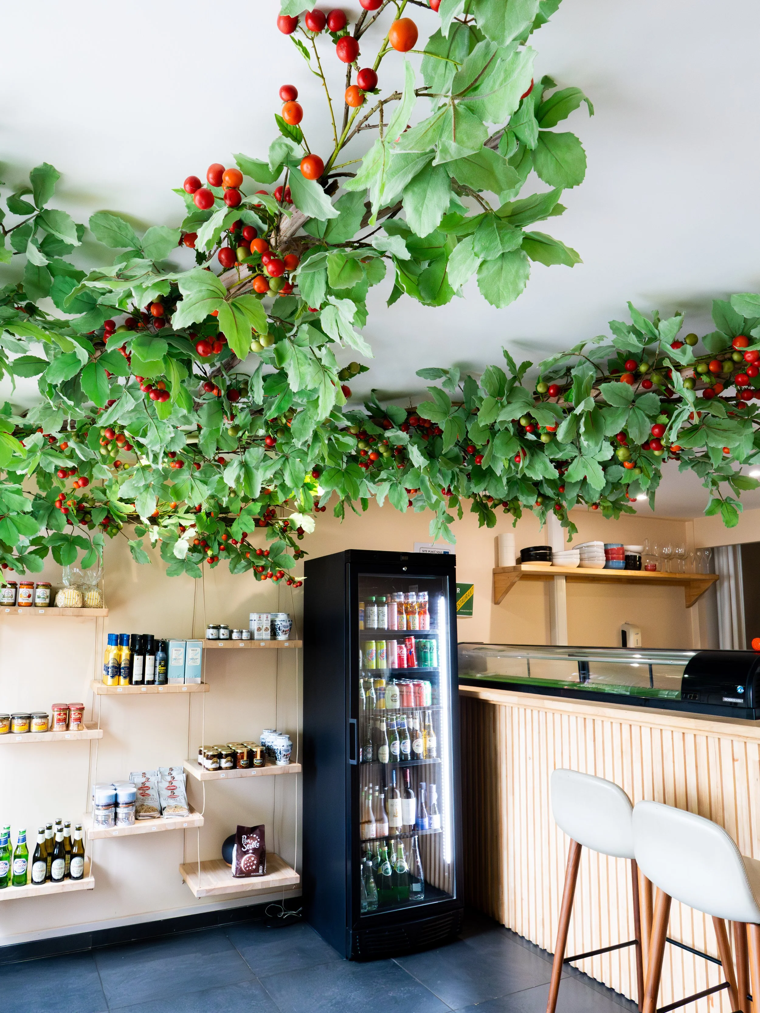 Intérieur d'une boutique ou café avec un plafond décoré de branches d'arbre avec des feuilles vertes et des baies rouges. Étagères avec des produits alimentaires en conserve, bouteilles et autres articles, et un réfrigérateur rempli de boissons. Quelques chaises blanches avec des jambes en bois sont visibles.