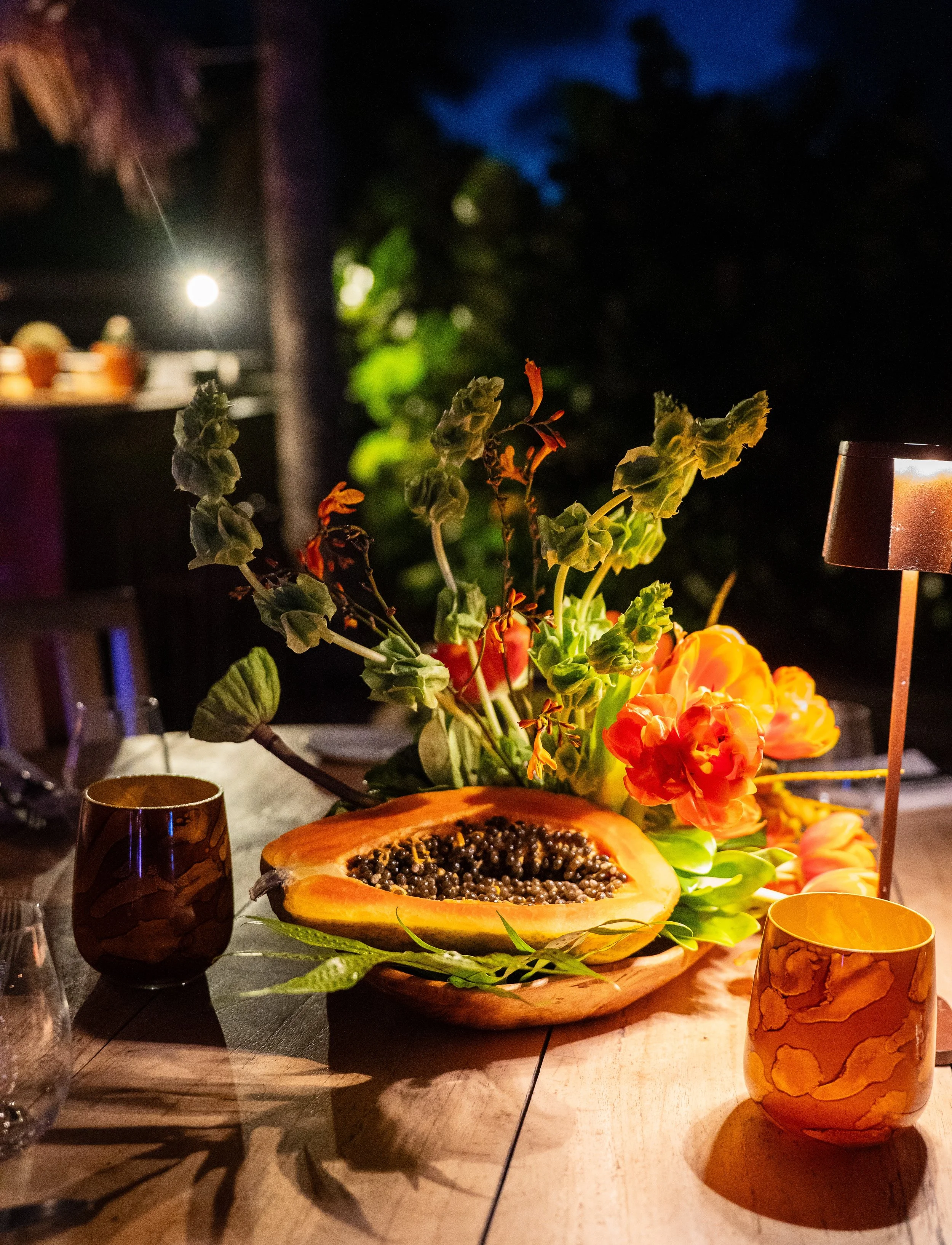 Un arrangement floral avec des fleurs orange, un demi-papayer avec des graines noires, une lampe à givenchy, et des verres en verre ornés de motifs amidon.