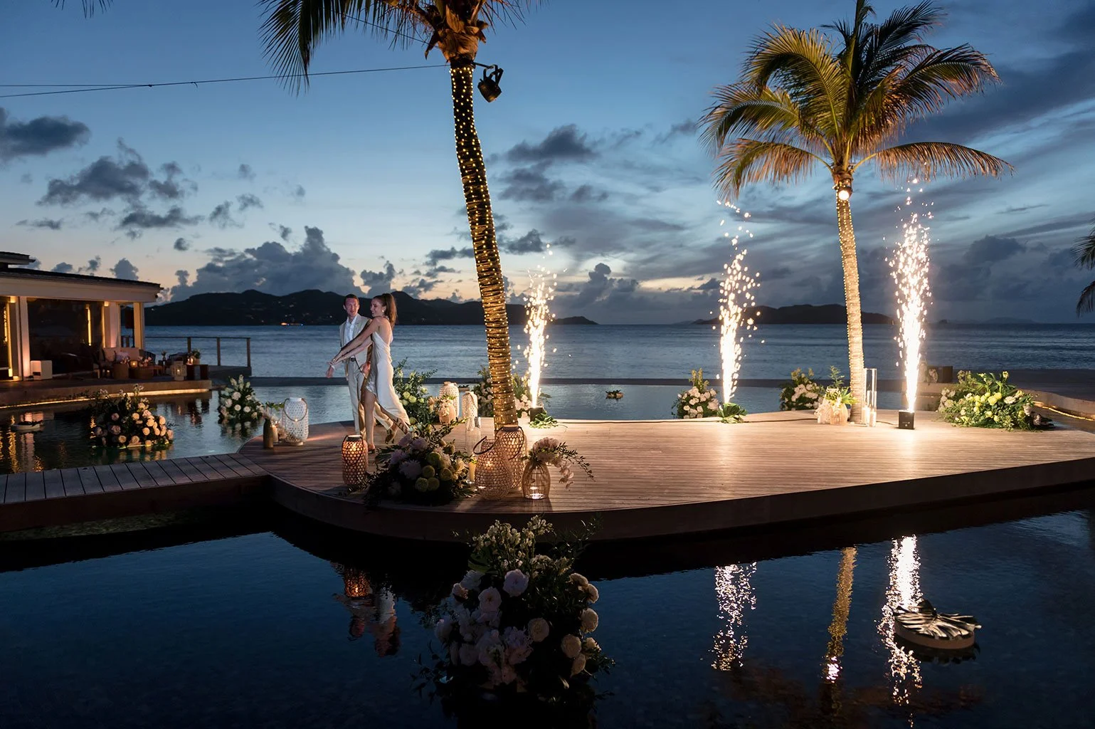 Une scène de mariage en plein air sur une terrasse en bois près de la mer, avec trois palmiers décorés de guirlandes lumineuses, des feux d'artifice et des fleurs accentuant l'ambiance nocturne romantique.