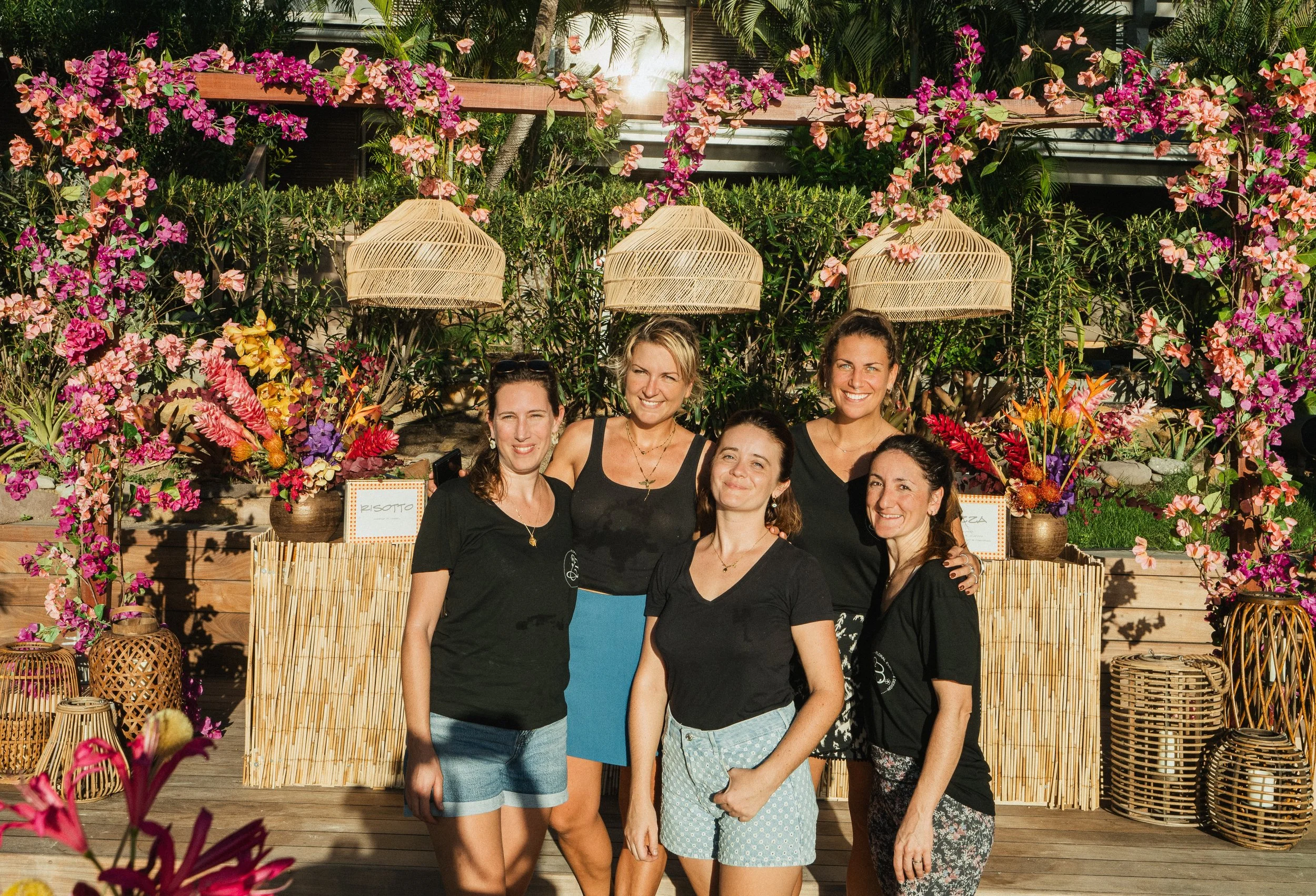 Groupe de femmes souriantes posant devant un fond de fleurs colorées comme des bougainvilliers dans un cadre extérieur