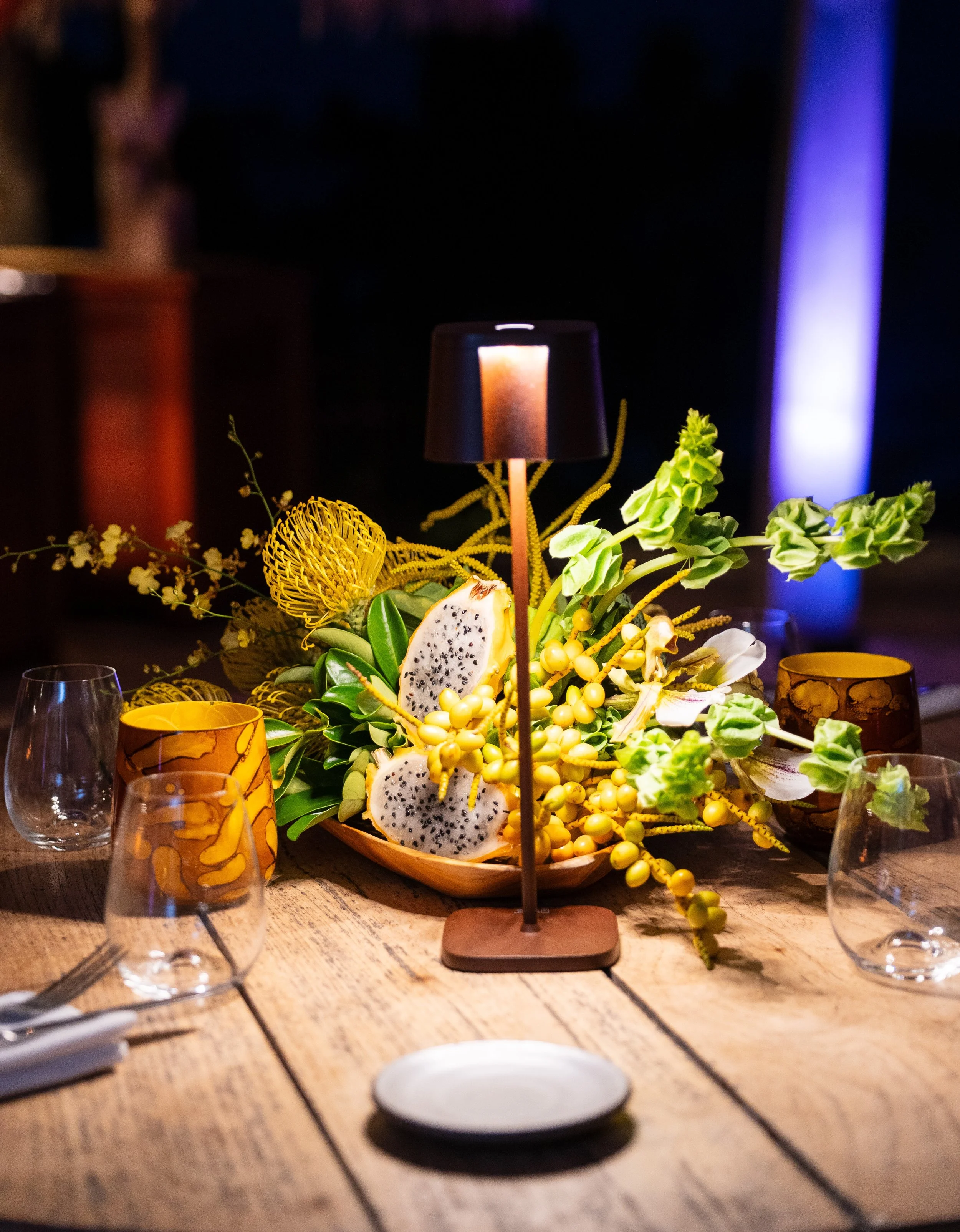 Un centre de table avec des fruits exotiques, des fleurs et des éléments décoratifs, éclairé par une lampe