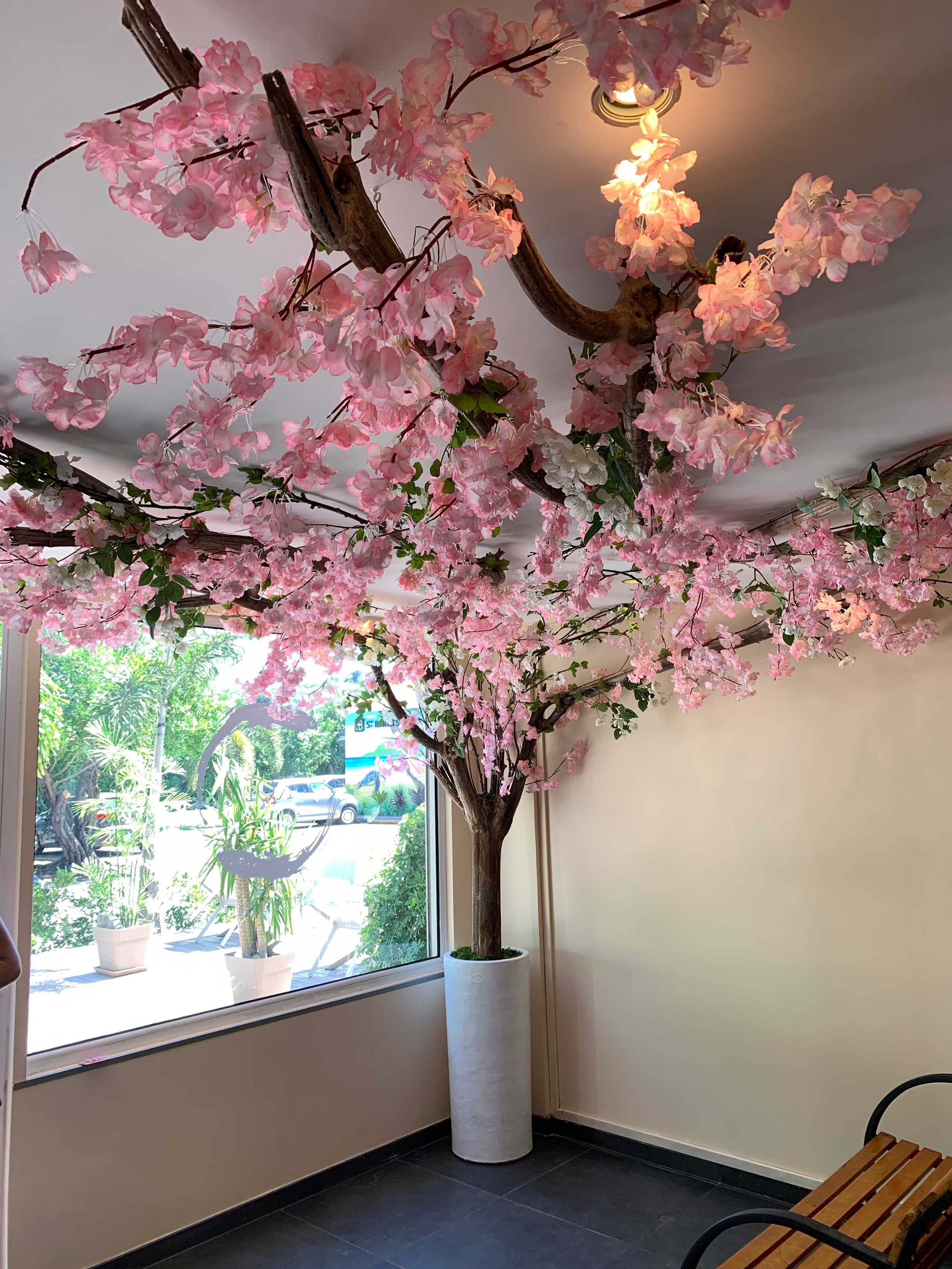 Indoor artificial tree with pink flowers in a white pot near a large window overlooking a garden, illuminated by soft ceiling light in St Barth.