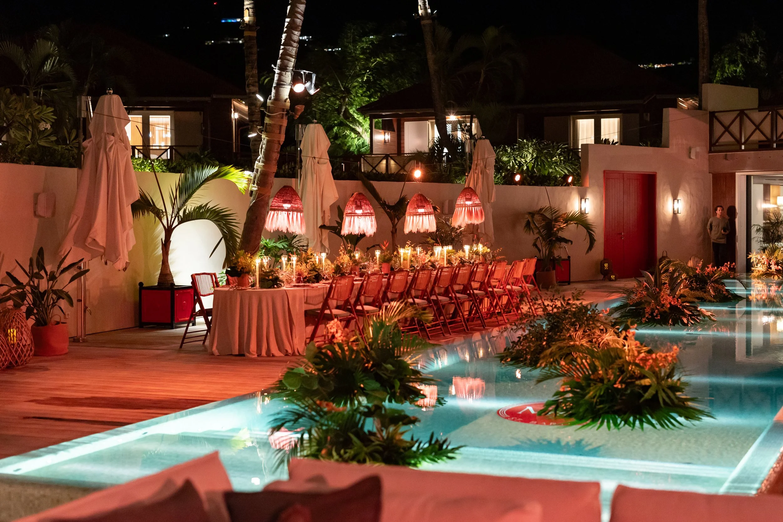 Décoration extérieure d'une soirée avec une table drapée, lampes en forme de parasol, plantes tropicales, et une piscine illuminée dans un environnement résidentiel la nuit.