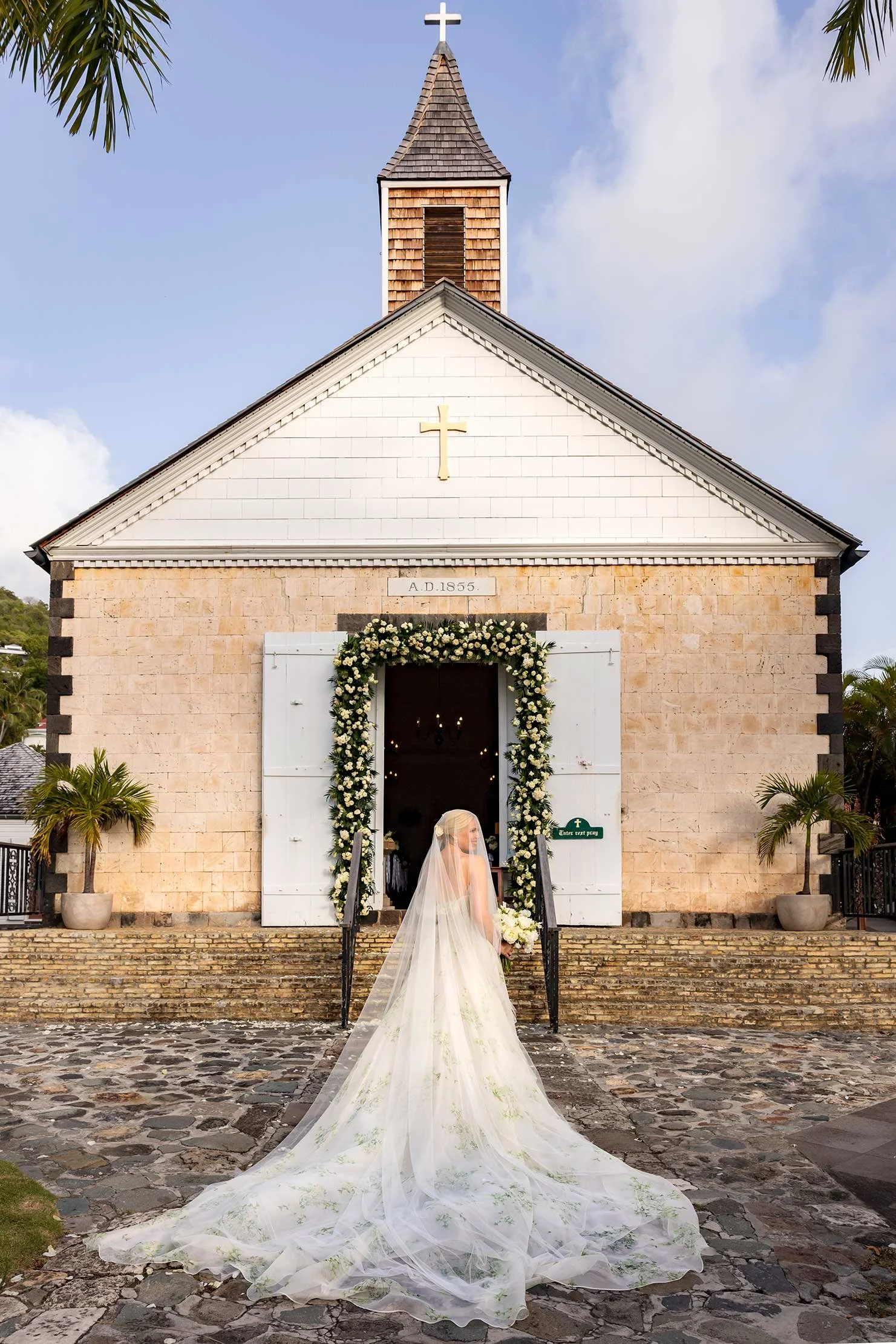 Elegant bride facing a white chapel adorned with cascading white floral arrangements for her wedding ceremony in St Barth.