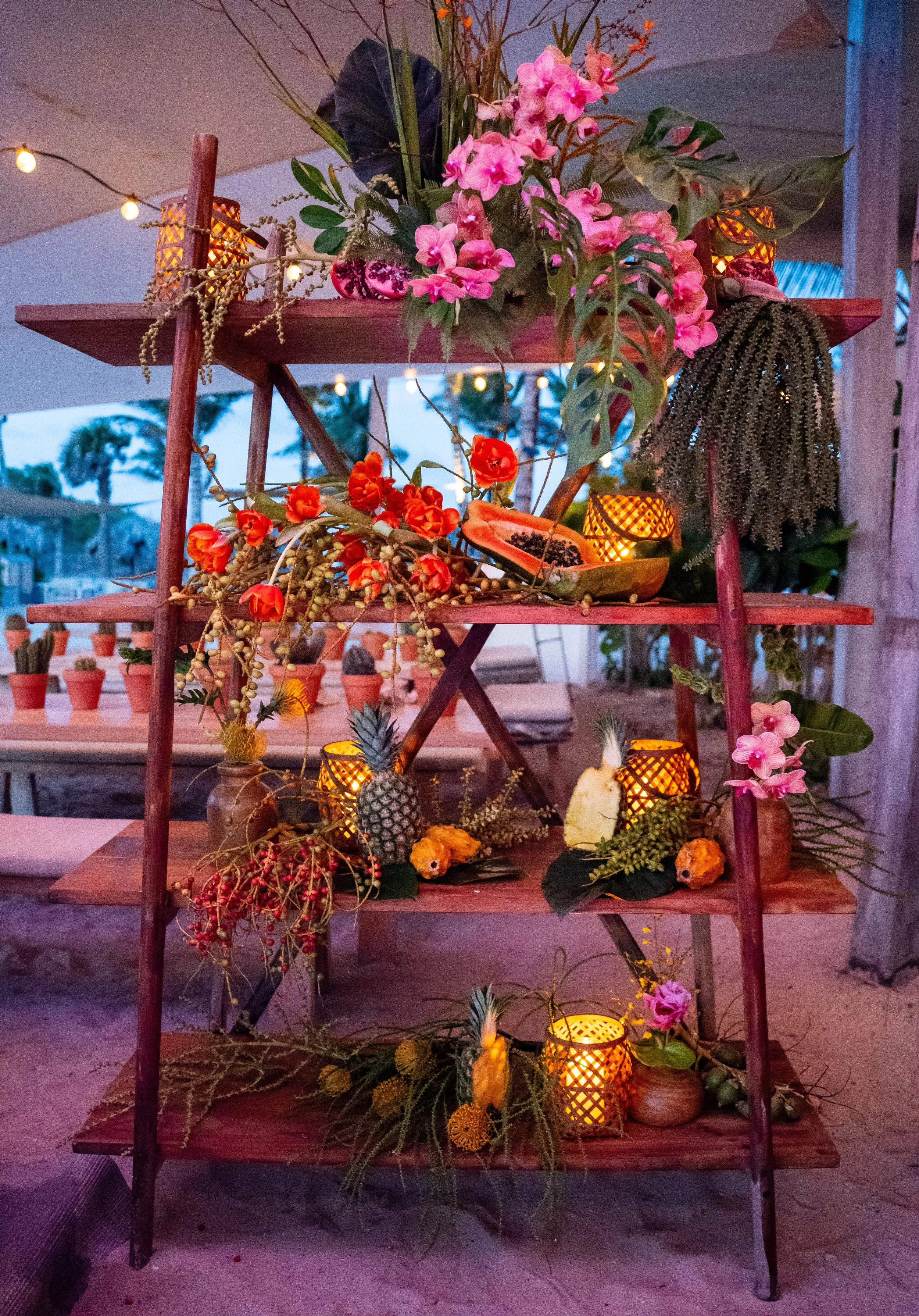 Disposition de décor tropical avec fleurs roses et oranges, ananas, papaye, papayer, et petits luminaires jaillissants, dans un cadre extérieur lors du crépuscule.