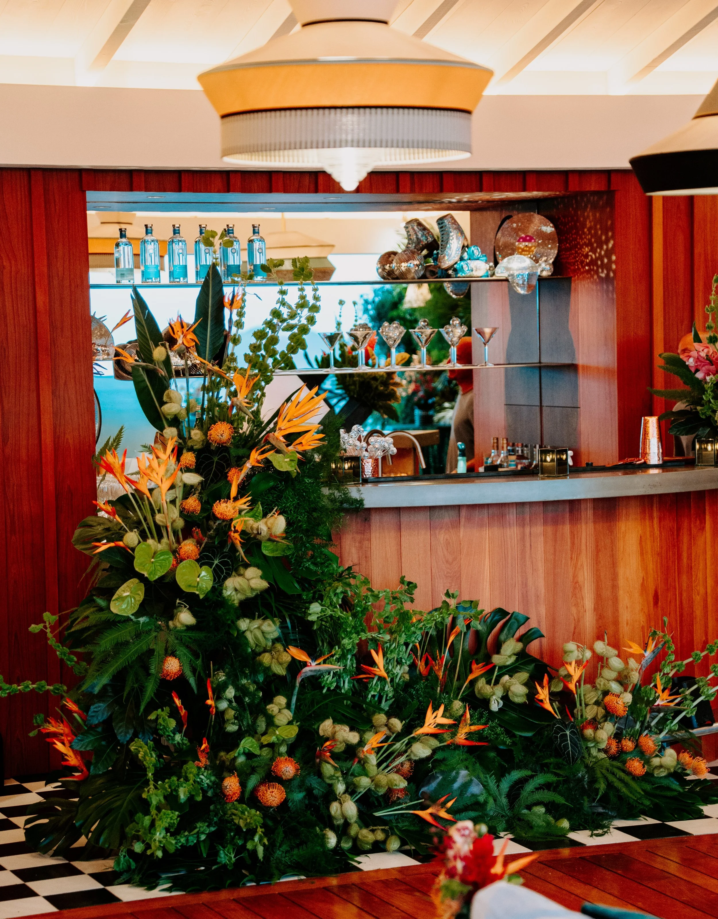 Une décoration florale élaborée avec de nombreuses fleurs colorées, placée devant un comptoir en bois avec des bouteilles d'eau alignées en arrière-plan.