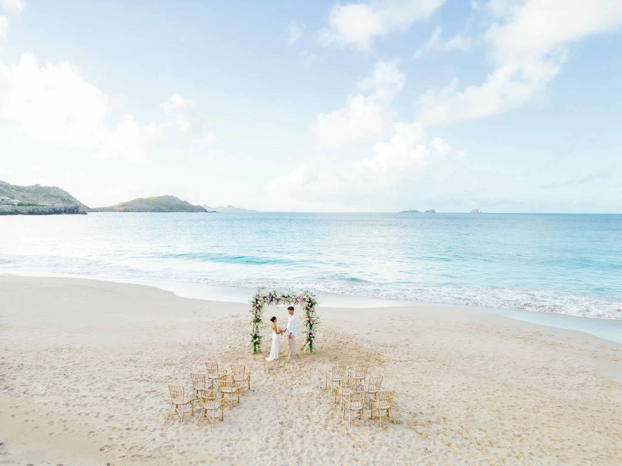 Romantic beach wedding in St Barth with a couple standing under a floral arch, surrounded by chairs arranged toward the ocean for a tropical ceremony.