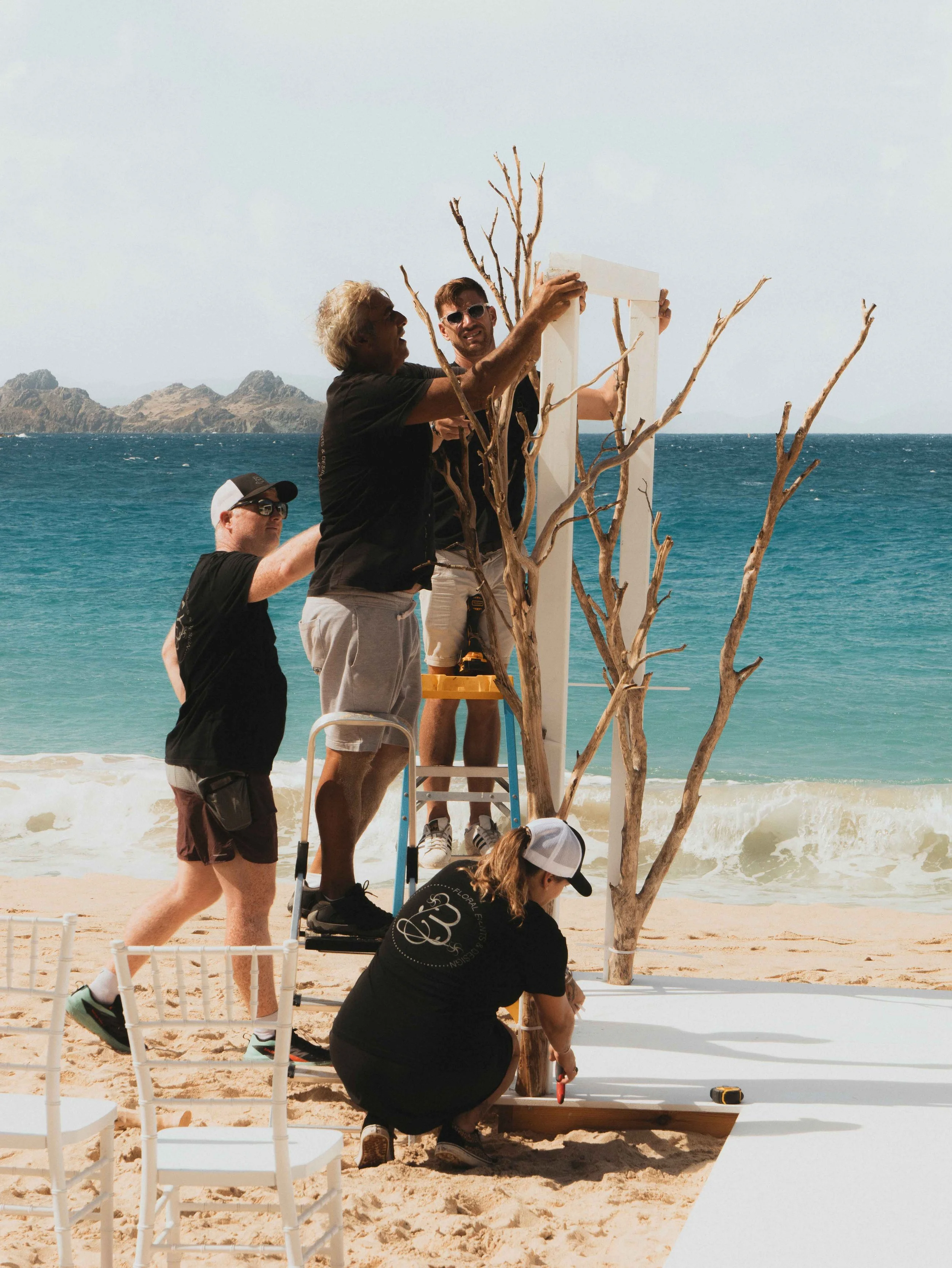 Production team building a beach wedding arch with driftwood and white panels on the sand in St Barth.