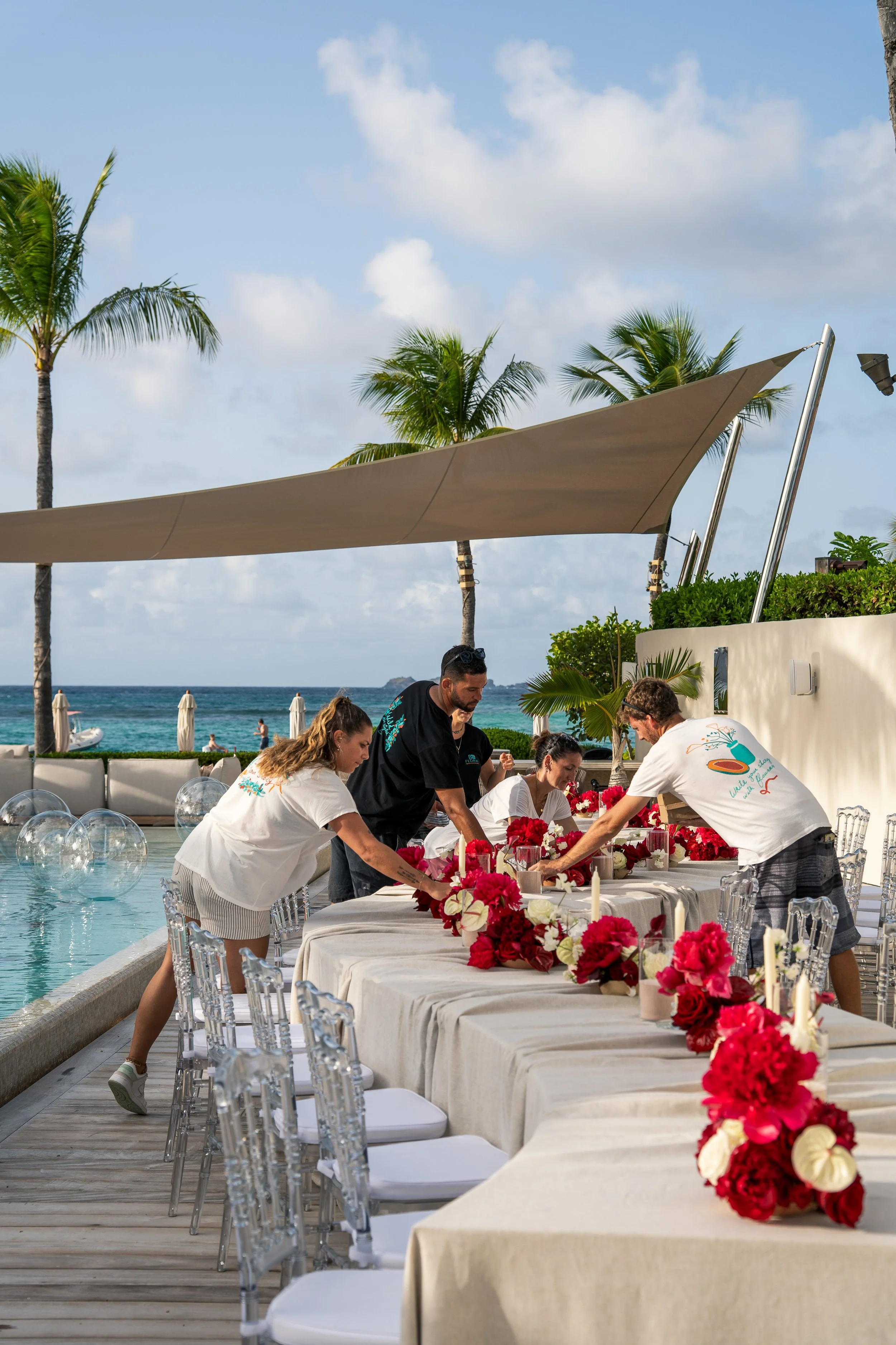 Un groupe de personnes prépare une décoration florale autour d'une longue table blanche, située près d'une piscine, avec vue sur la mer, des palmiers, un parasol beige, et un ciel partiellement nuageux en arrière-plan.