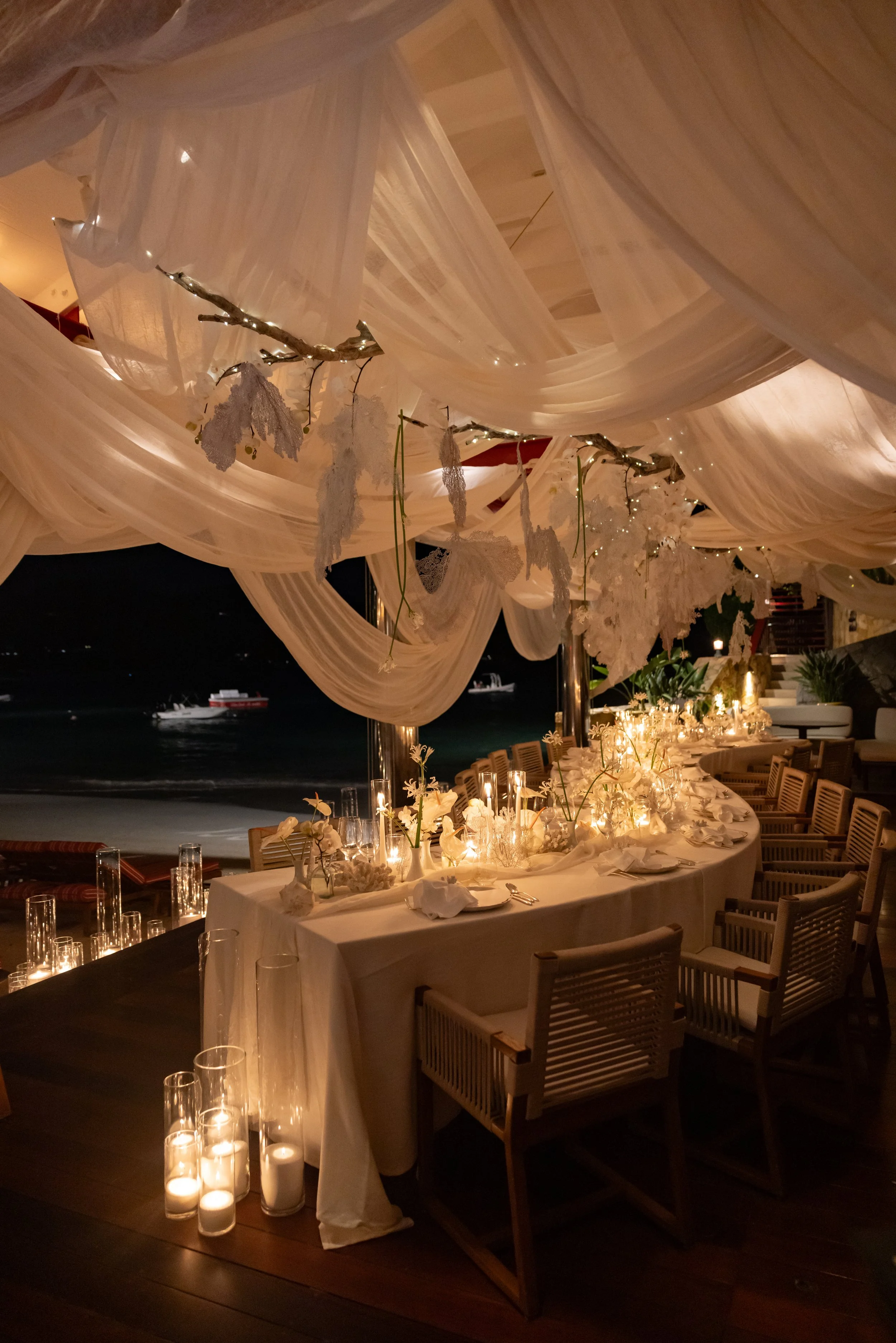Dîner de mariage en plein air avec des draperies blanches suspendues et des bougies sur la table, vue nocturne sur la mer avec un bateau en arrière-plan.