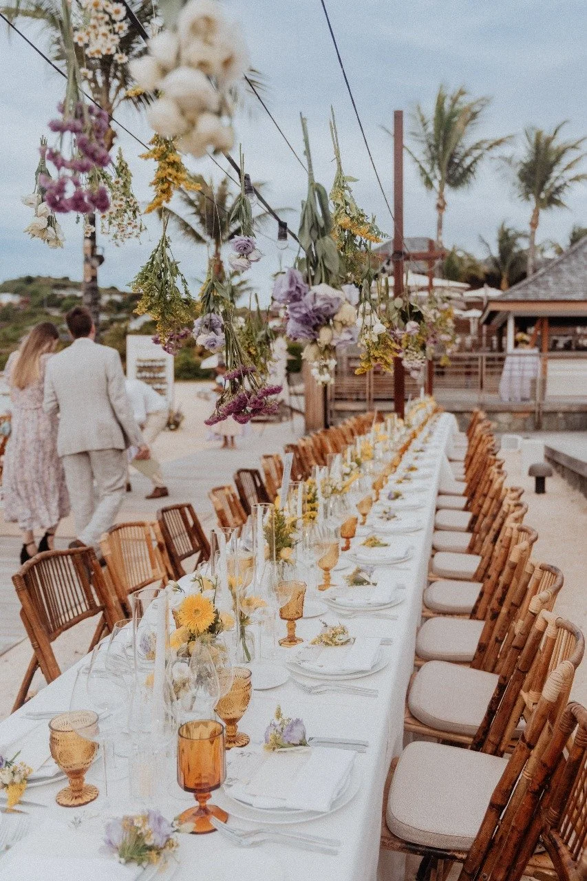 Beach wedding dinner setup with a long table decorated with yellow flowers, amber glassware, and bamboo chairs overlooking the sea in St Barth.