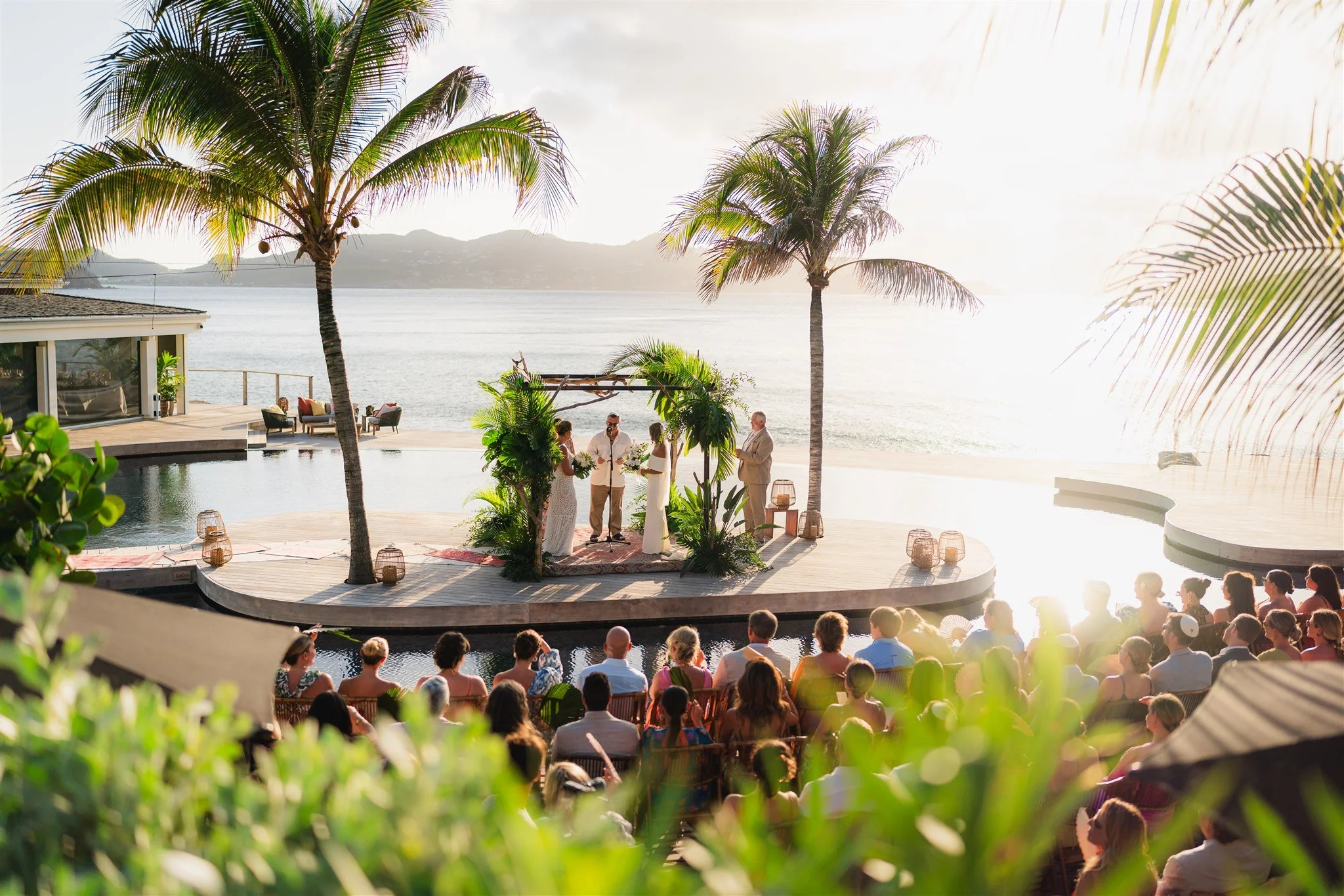 Cérémonie de mariage en plein air sur une plateforme en bois près de la mer, entourée de palmiers, avec des invités assis regardant la cérémonie et la mer en arrière-plan au lever ou coucher du soleil.