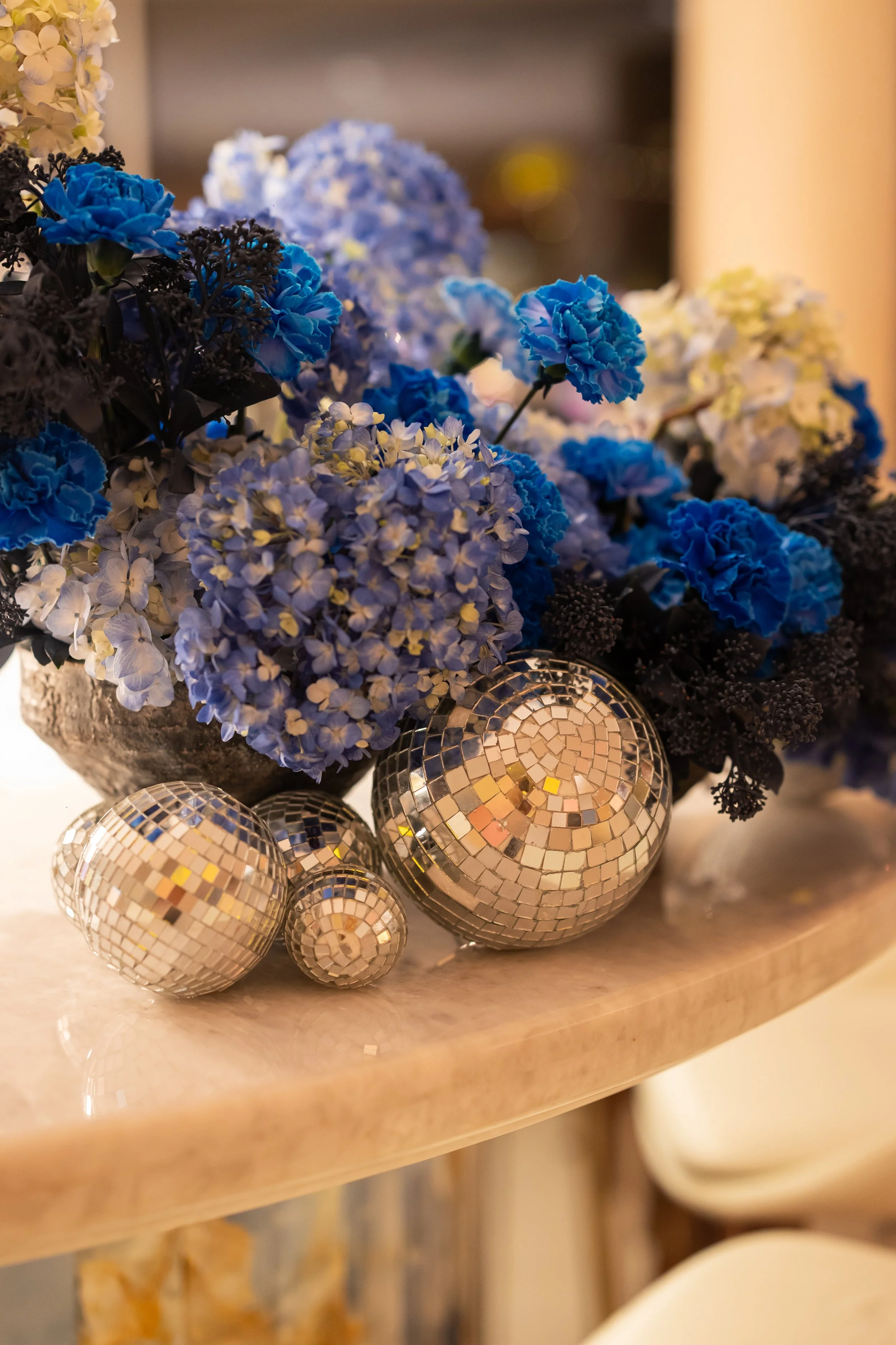 Un arrangement floral composé de fleurs bleues et blanches, entouré de boules décoratives en mosaïque argentée, placé sur une surface en pierre ou en marbre.