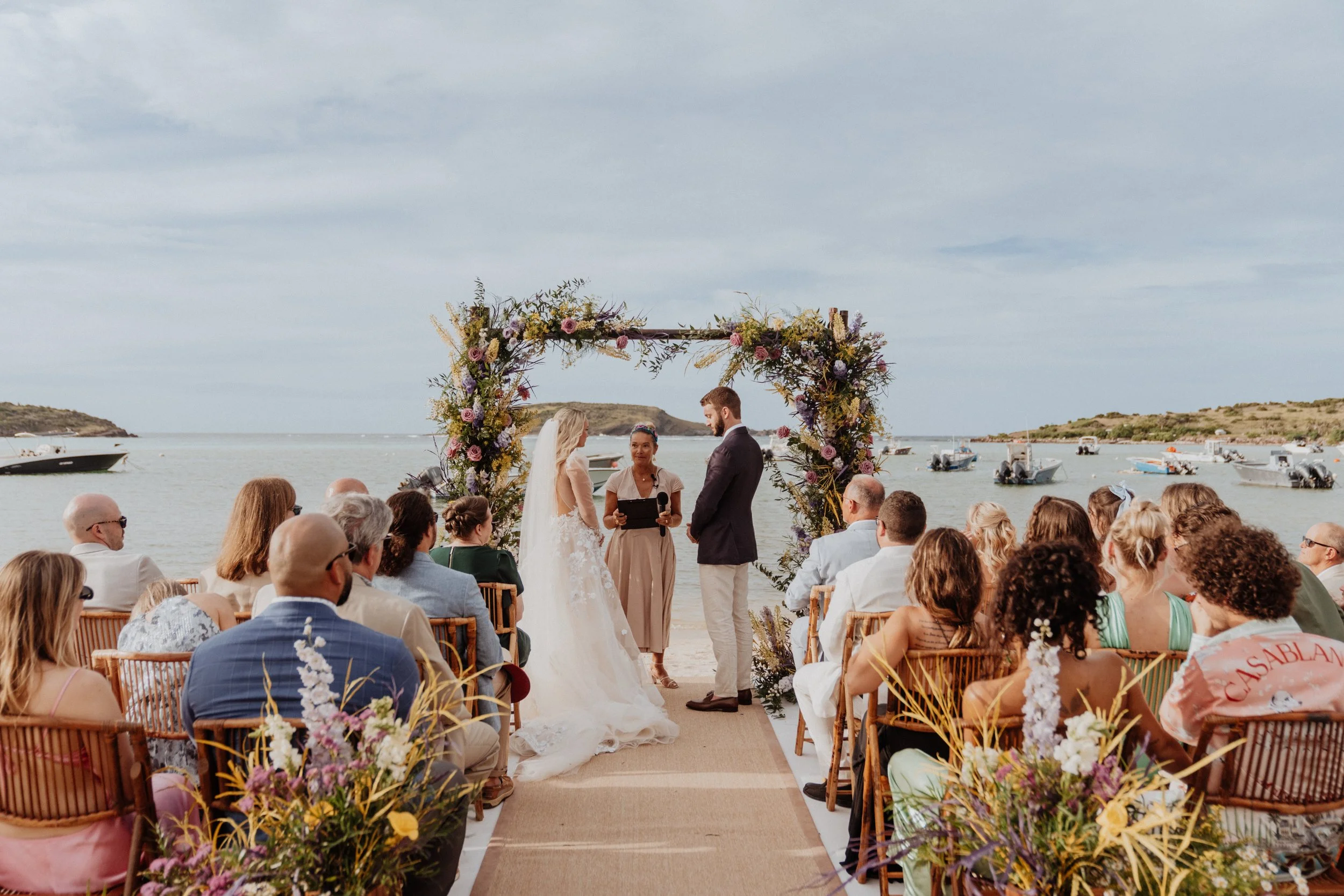 Romantic beach wedding ceremony in St Barth featuring a floral arch, bride and groom, guests, and ocean views with boats in the background.