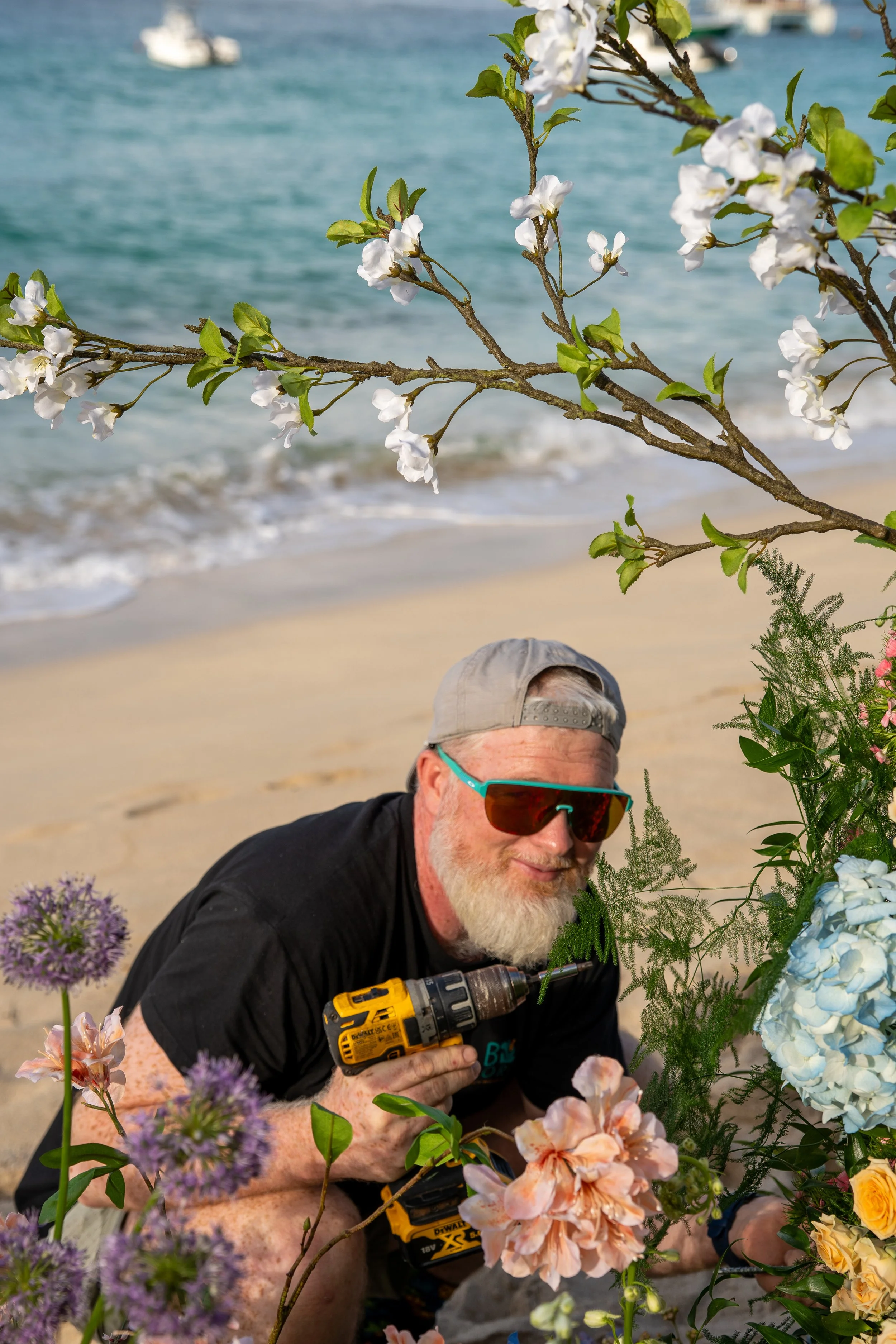 Un homme âgé portant des lunettes de soleil, un chapeau gris et une veste noire, tenant une perceuse sans fil jaune, est assis sur la plage parmi des fleurs colorées, avec la mer en arrière-plan.