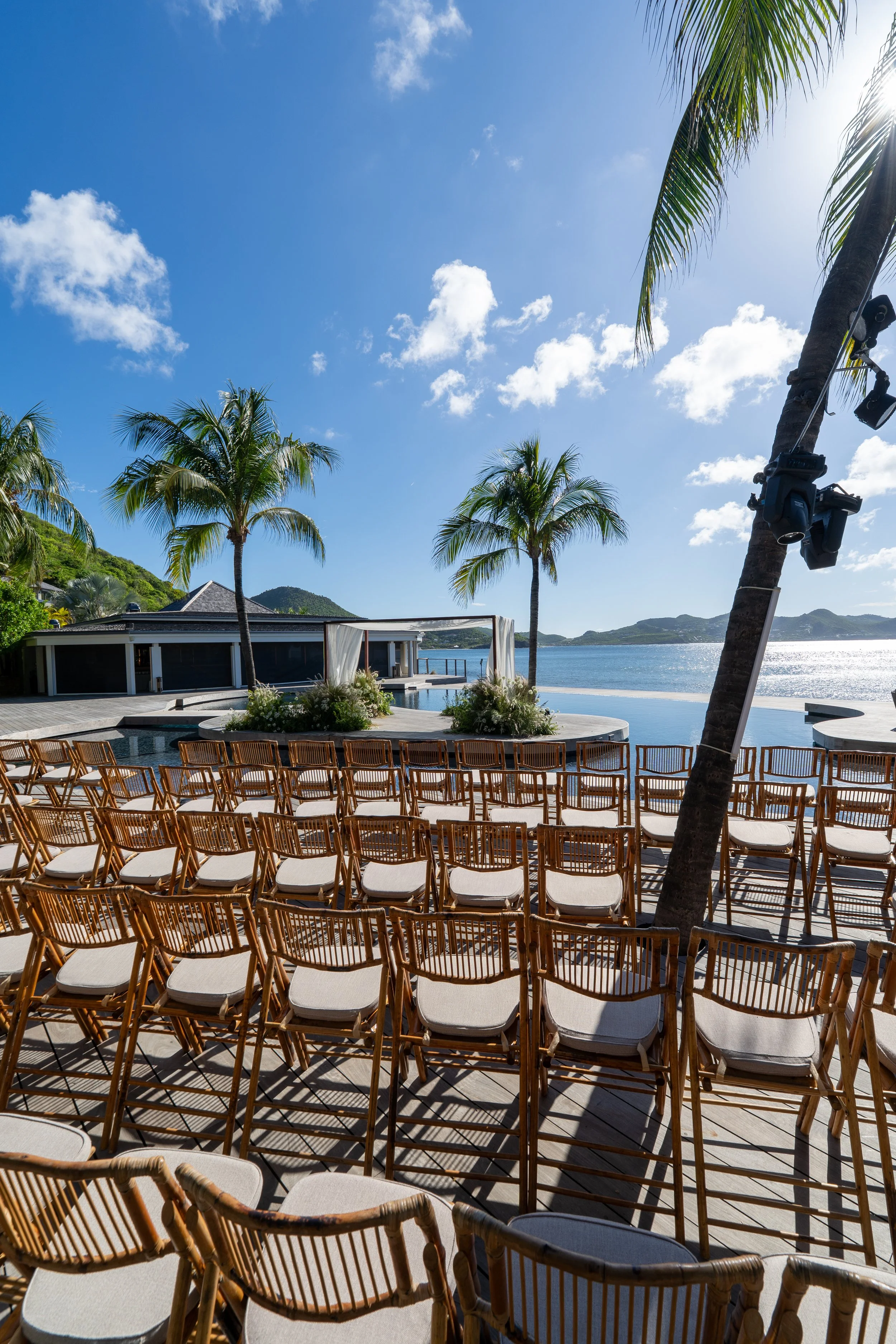 Une scène d'une cérémonie en plein air près de la mer avec des chaises en bois, des palmiers, une pergola blanche et un fond de montagnes sous un ciel bleu avec quelques nuages.
