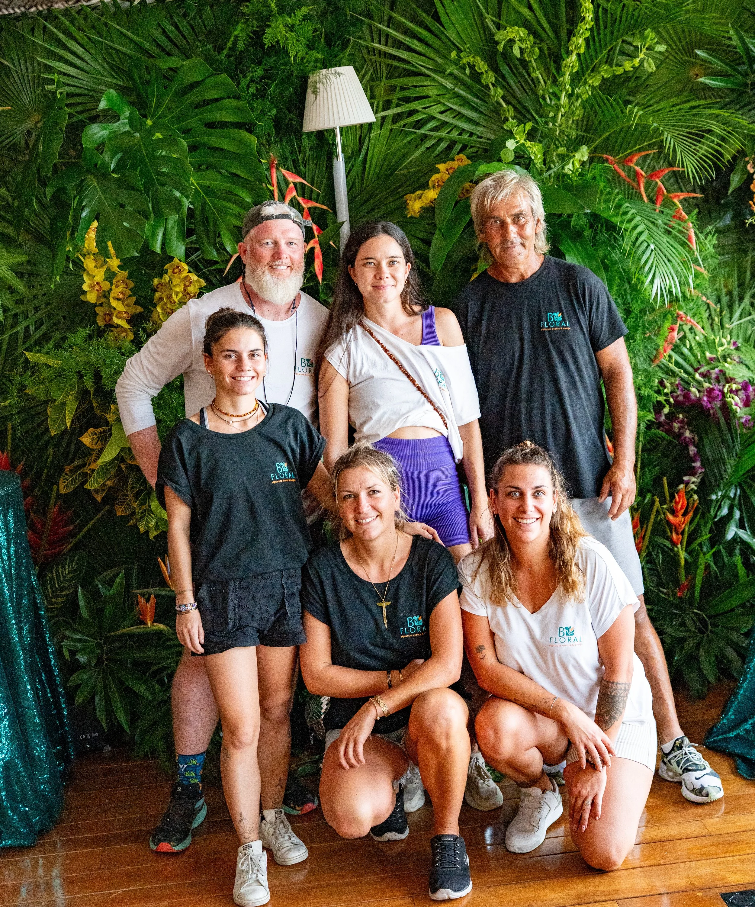 Groupe de neuf personnes posant devant un mur de feuillages verts et de fleurs tropicales, certains assis et d'autres debout, lors d'un événement ou d'une réunion.