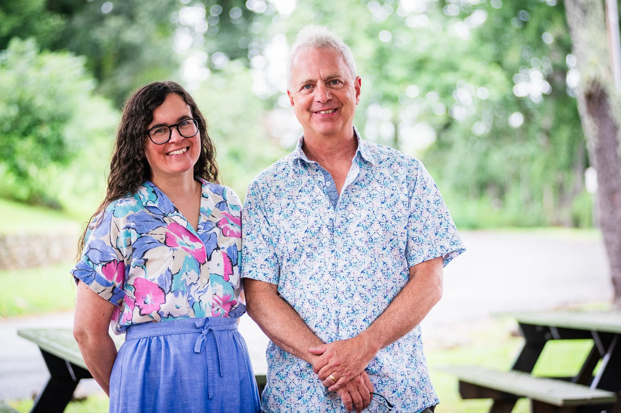 A woman with long curly dark hair and glasses wearing a colorful floral shirt and blue high-waisted pants, standing next to a man with short gray hair wearing a white floral shirt, outdoors in a park with green trees in the background.