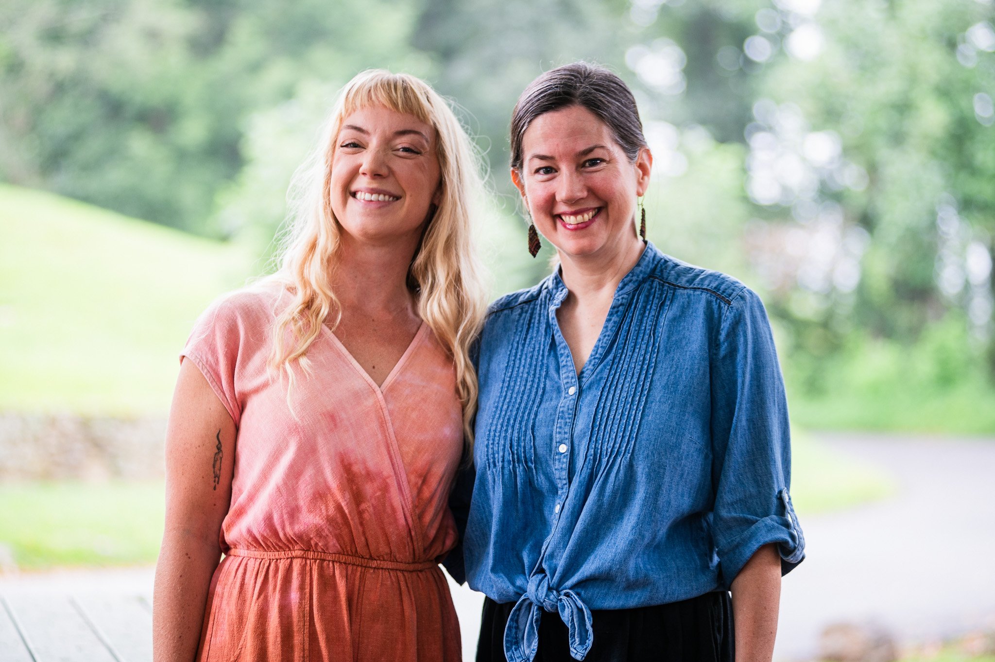 Two women standing outdoors, smiling with greenery in the background.