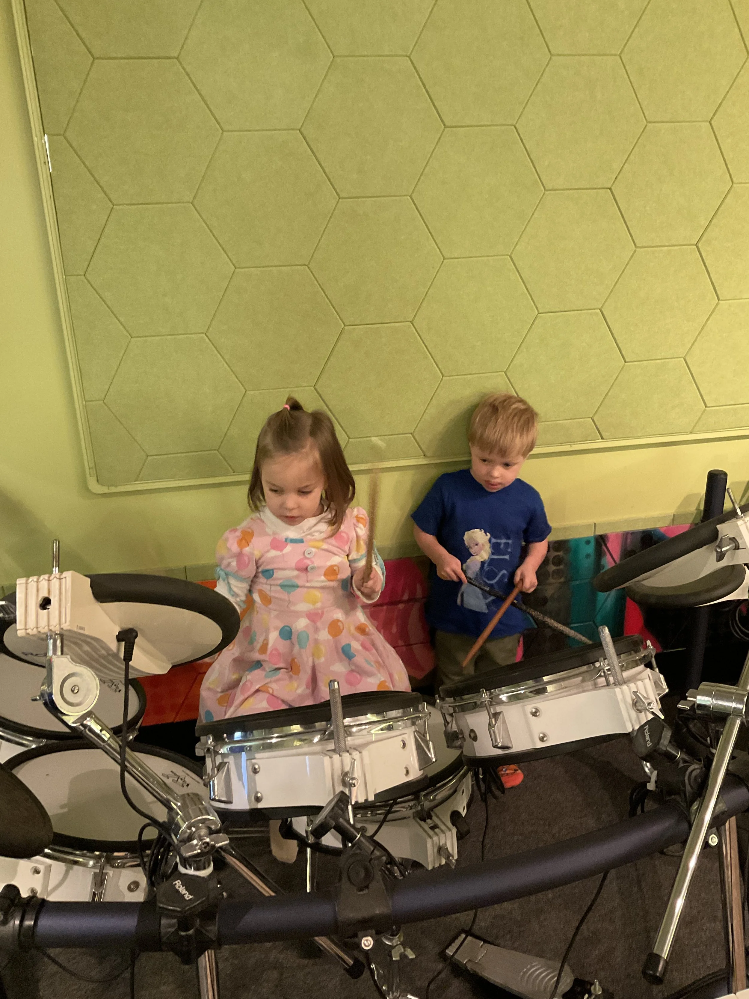 Two young children, a girl and a boy, are playing electronic drums in a room with yellow walls and a hexagonal pattern on the wall behind them.