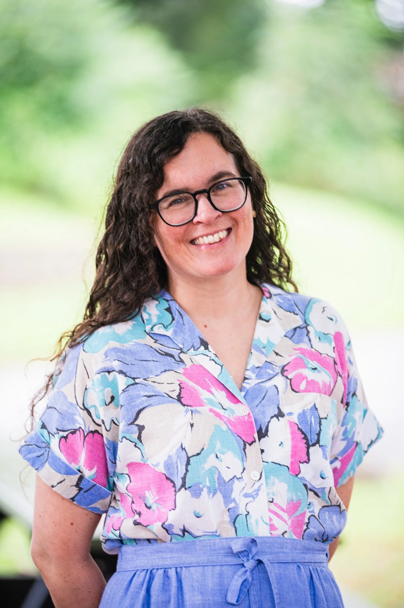 A woman with curly dark hair, glasses, and a bright smile stands outdoors in a park with green trees in the background.
