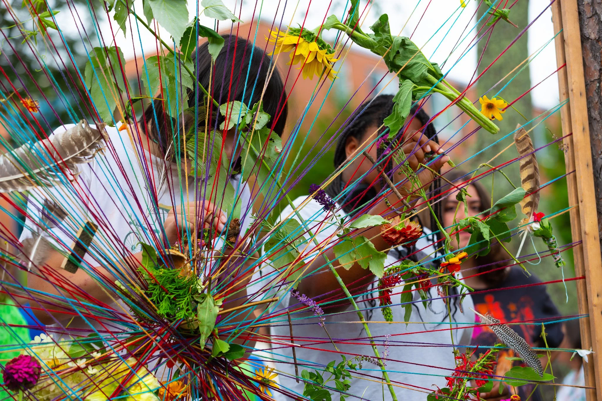 People participating in a yarn weaving activity, weaving flowers and leaves into a colorful web of strings on a wooden frame.