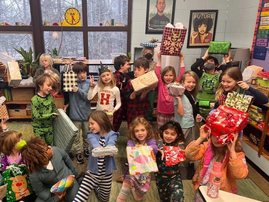 Children in pajamas and cozy clothes exchanging and holding presents in a classroom decorated for Christmas.