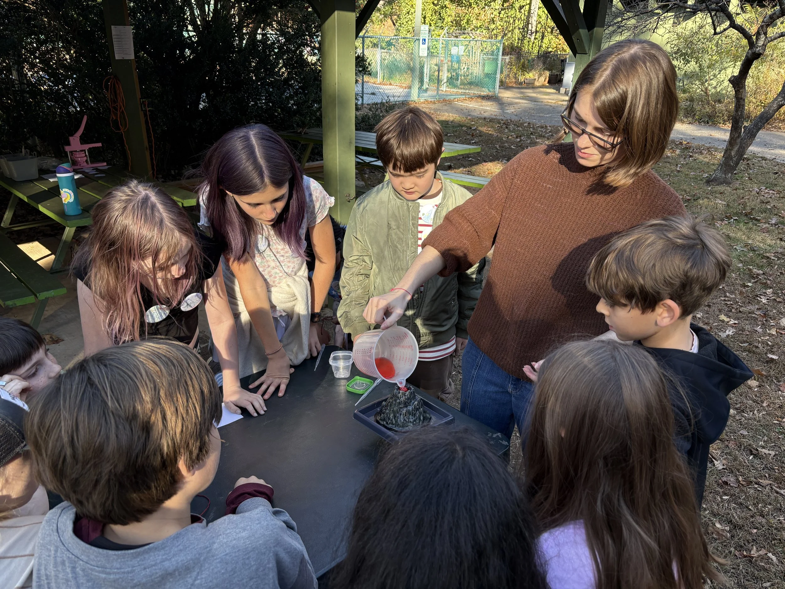 A group of children gathered around an outdoor table watching a woman pour red liquid onto a volcano model during a science demonstration.