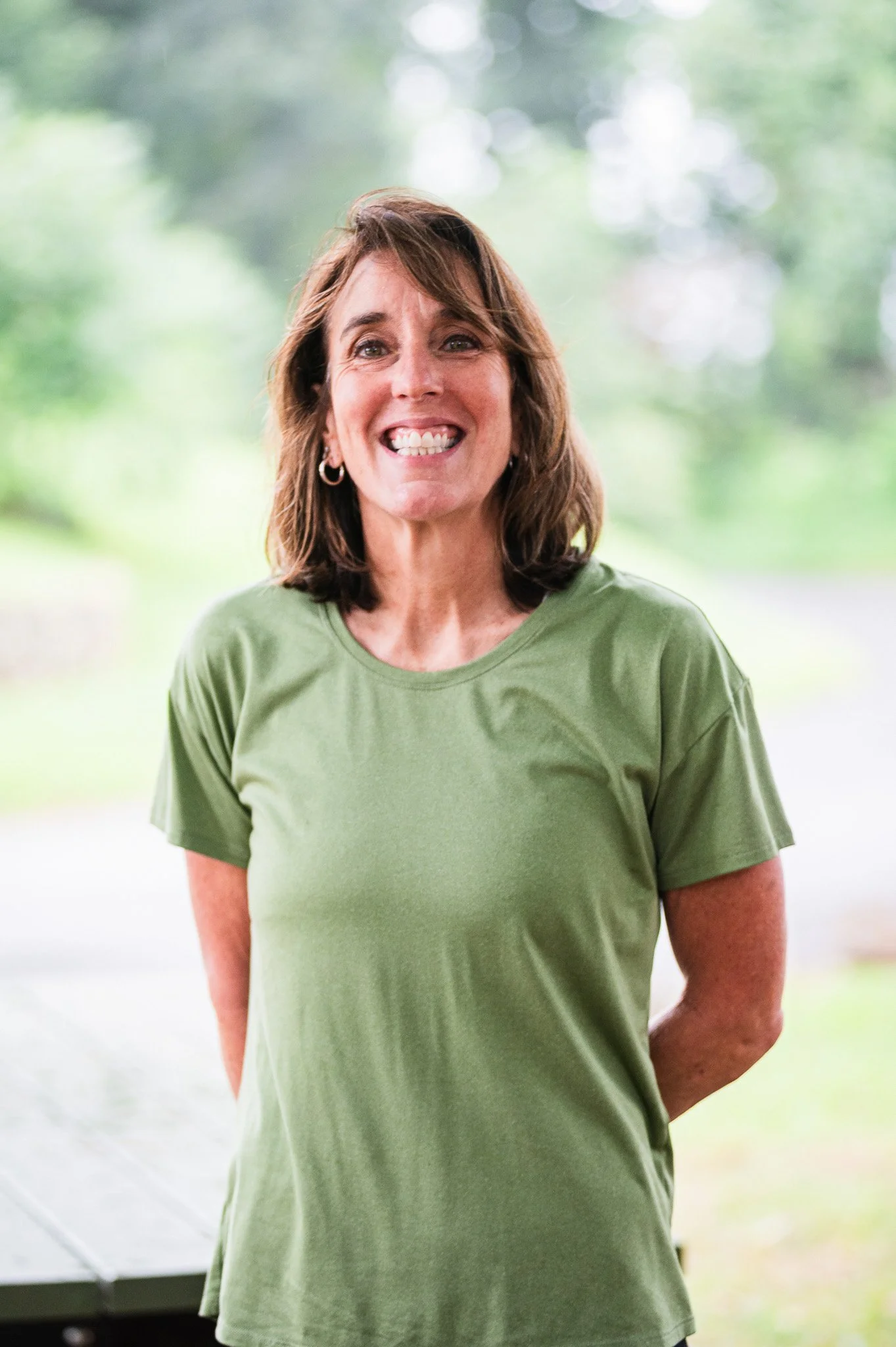 A woman with shoulder-length brown hair smiling, wearing a green T-shirt, and standing outdoors with green trees in the background.