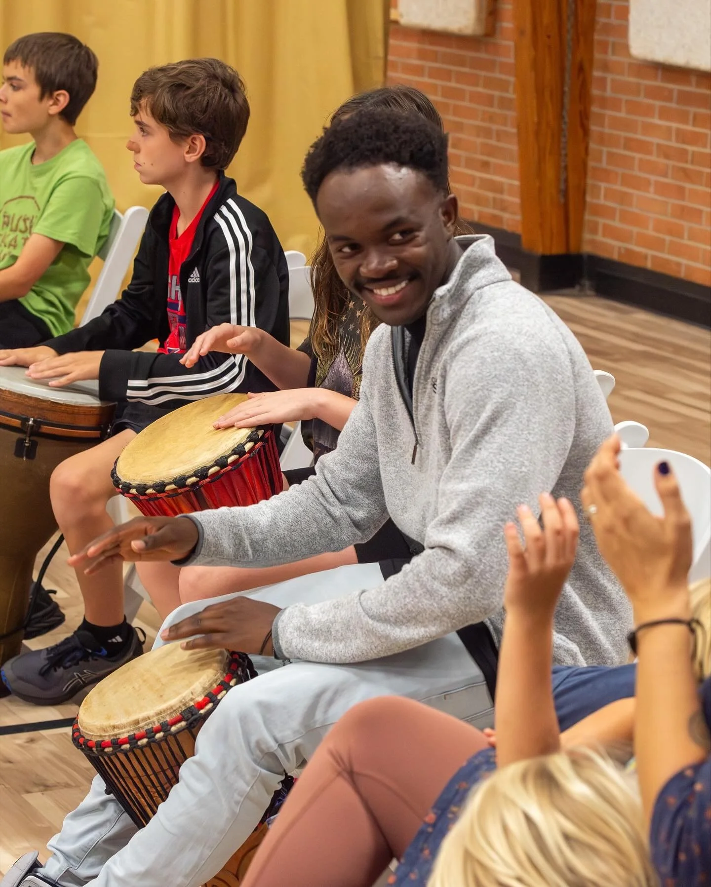 A group of people sitting indoors, playing drums and clapping. One man in a gray jacket smiling at the camera, surrounded by children playing drums.