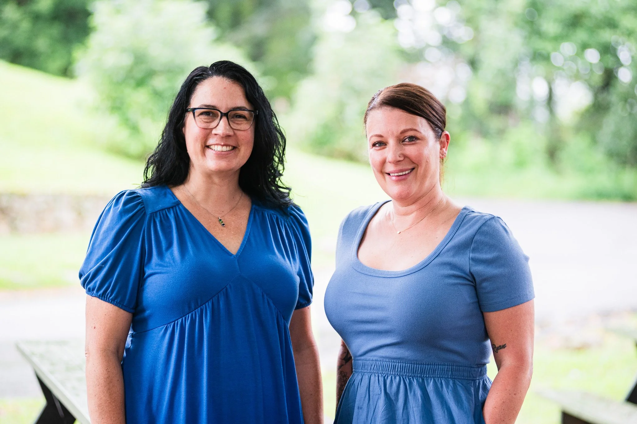 Two women standing outdoors, smiling, with trees and greenery in the background. One woman has black hair, glasses, and is wearing a blue dress, while the other has brown hair and is wearing a blue top.