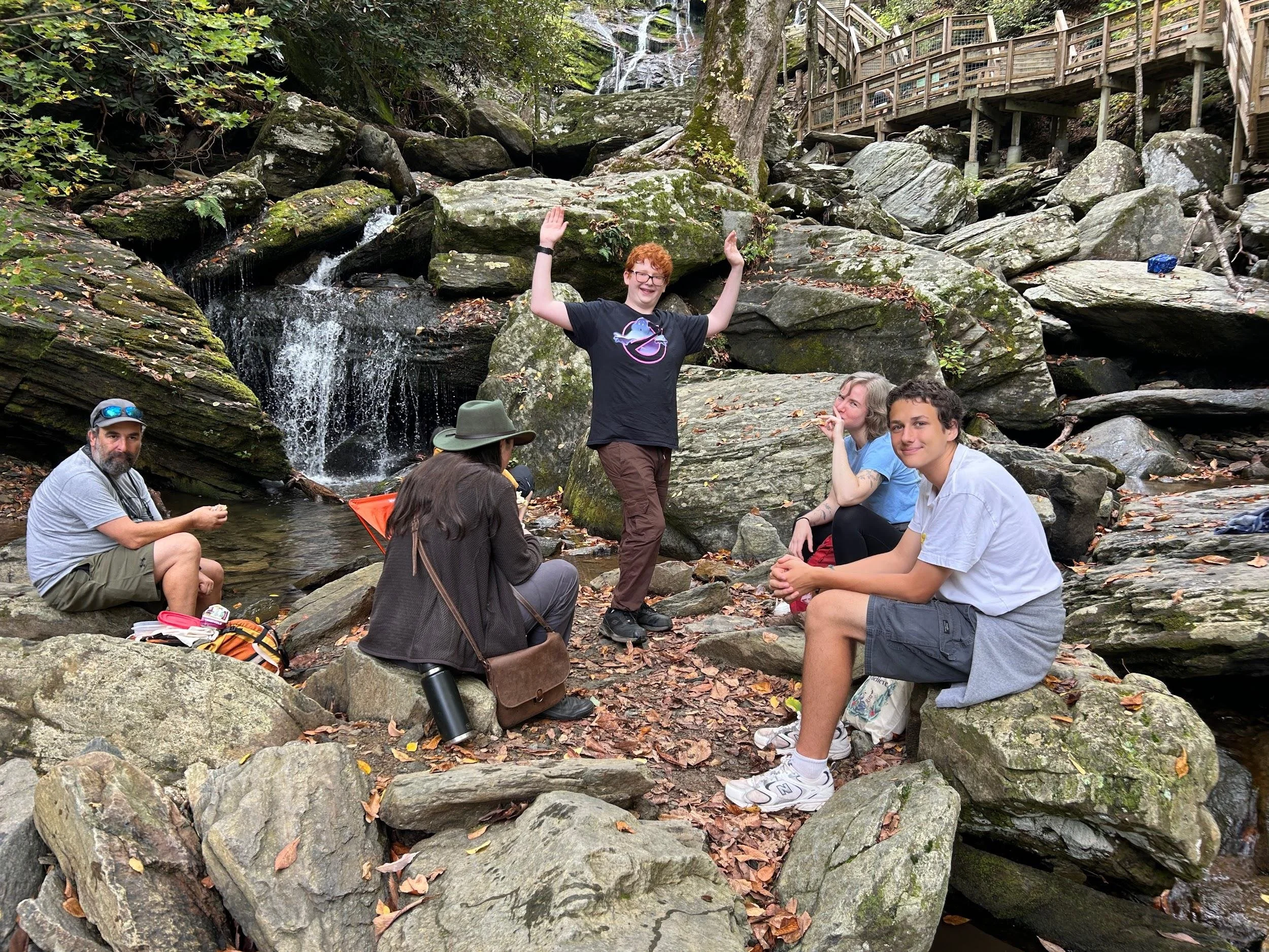 Group of five people sitting and standing on rocks near a small waterfall in a wooded area.