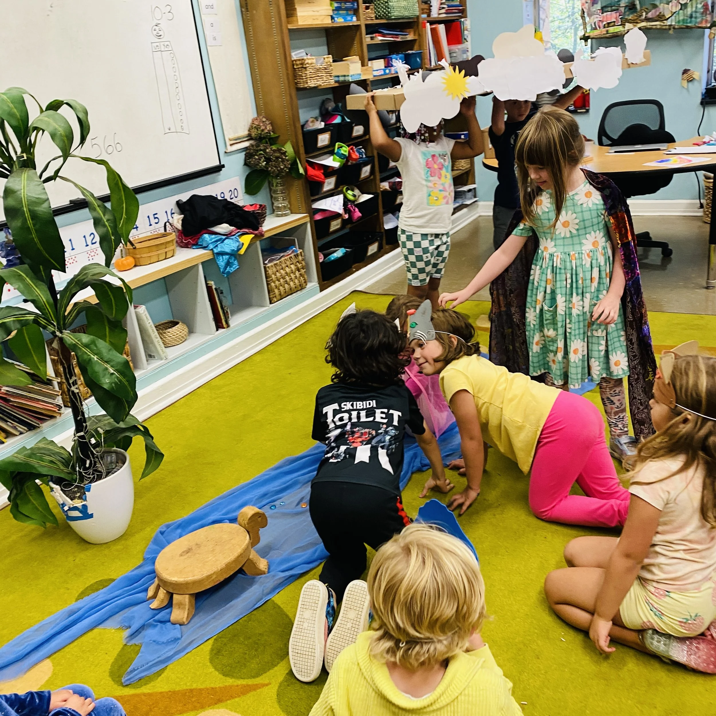 Children in a classroom engaged in a play acting scene. Some children are on the floor, with two children in costumes crouching and smiling at each other. Others are standing and holding props or making a cloud shaped hat. The classroom has educational materials, bookshelves, and a whiteboard.