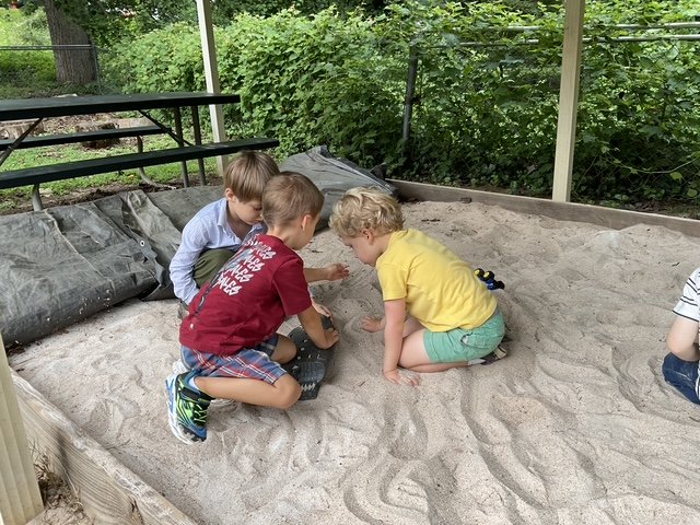 Three children playing in a sandbox on a screened porch, surrounded by green trees and bushes.