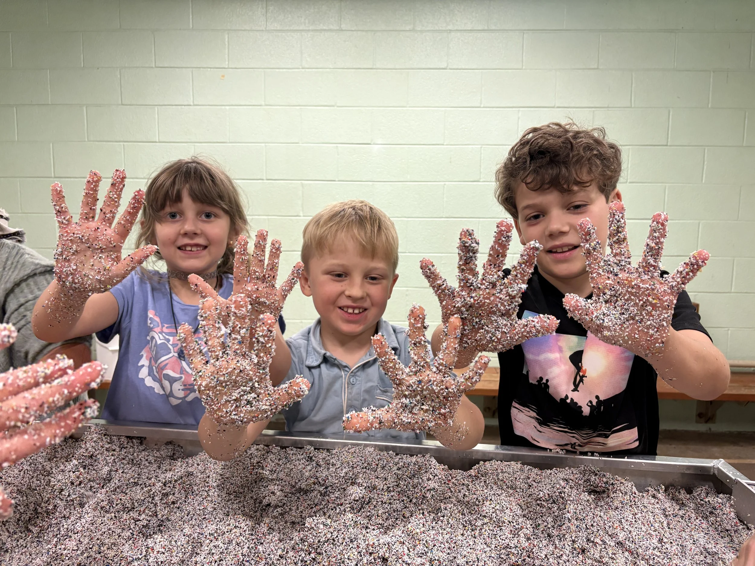 Three children with glitter-covered hands smiling and showing their hands at a sensory table filled with colorful tiny beads or grains.