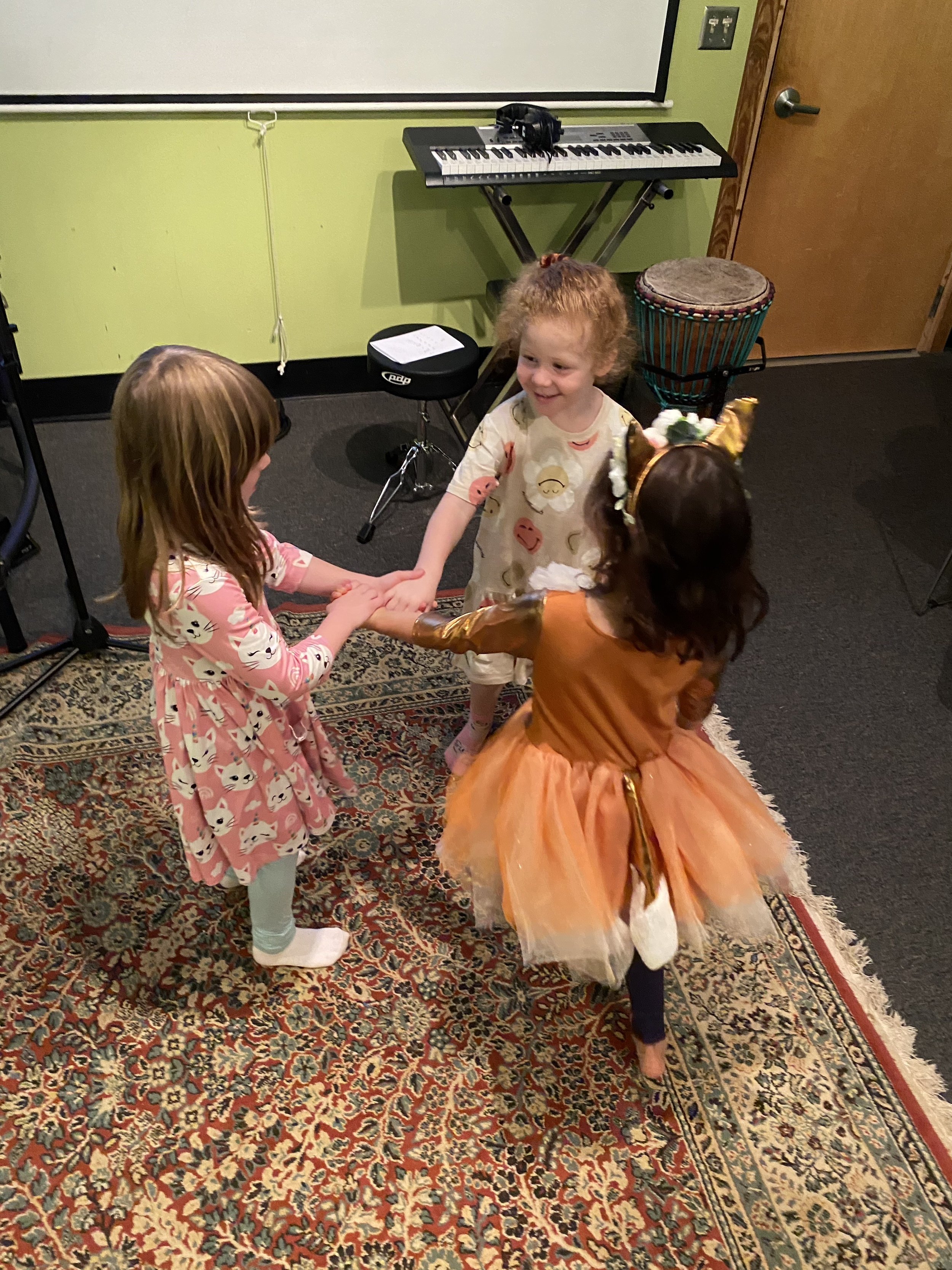 Three young girls are holding hands and dancing in a circle in a room with a patterned carpet. One girl is dressed in a pegasus costume with a tutu, while another wears a pink dress with cat faces. The third girl has curly hair and is wearing pajama-