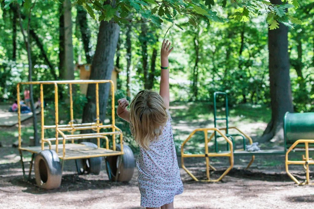 A young girl with blonde hair wearing a floral dress reaches up to touch tree leaves in a wooded playground.