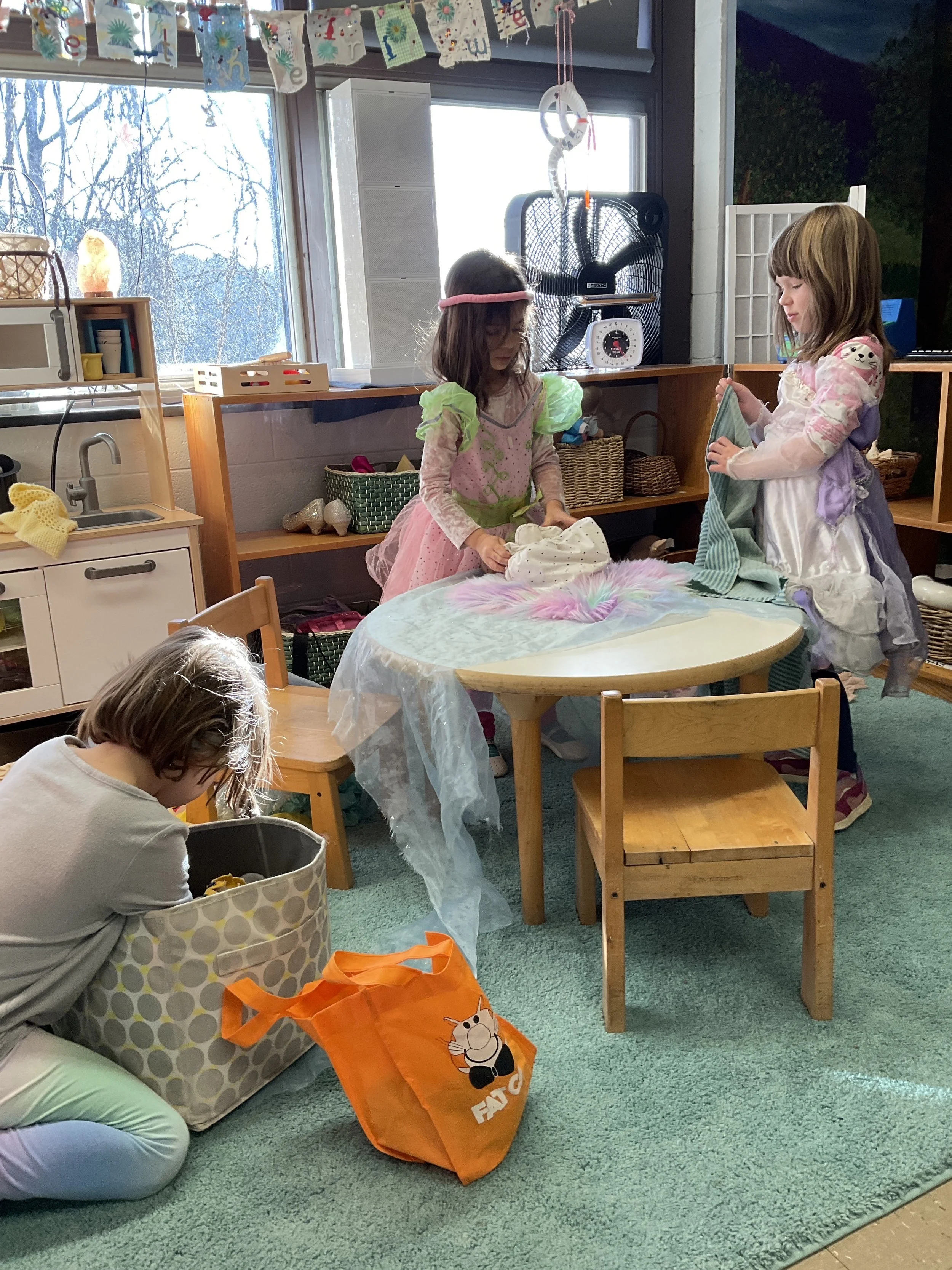 Two children dressed as princesses are playing with laundry on a table, while a third child is organizing items in a large storage container nearby in a playroom with a window and decorations.