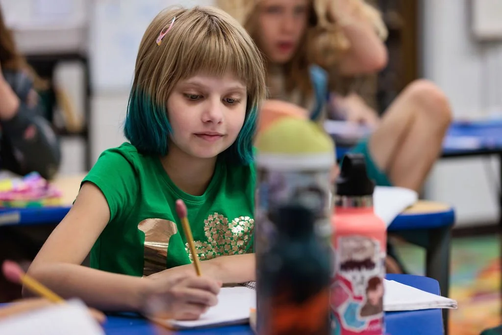 an elementary student with dyed hair focusing on her writing and smiling