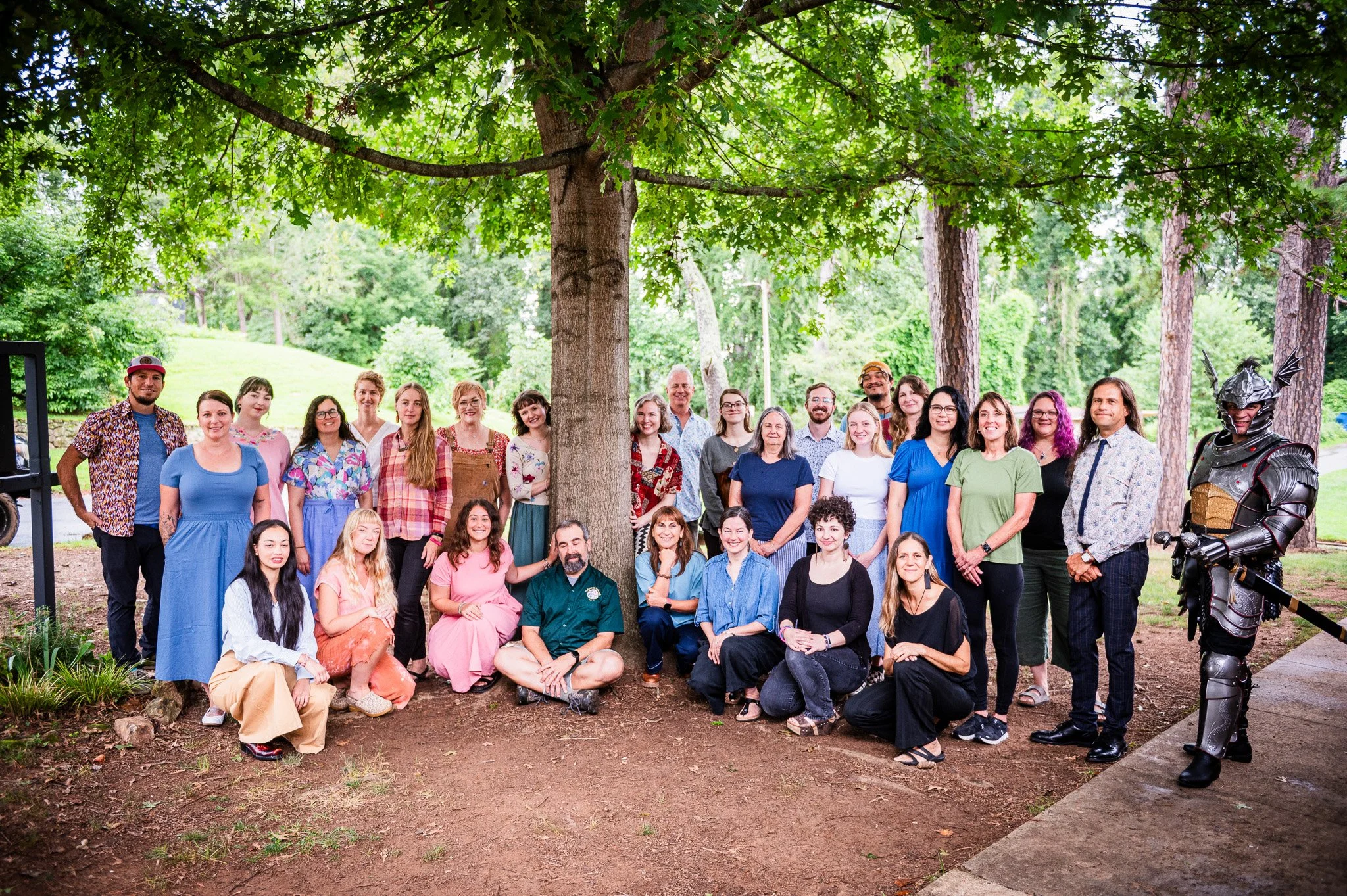 Group of diverse people standing and kneeling outdoors beneath a large tree during daytime, with a man in armor on the right side.