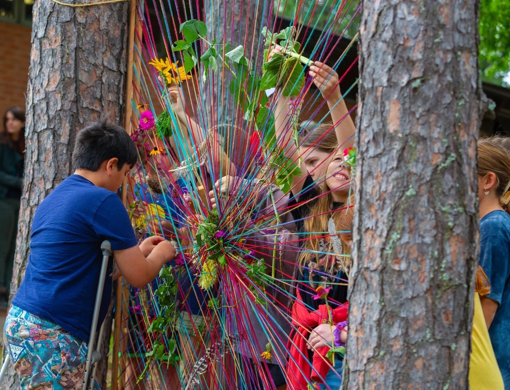 Group of young people and children creating a colorful yarn web among trees outdoors.