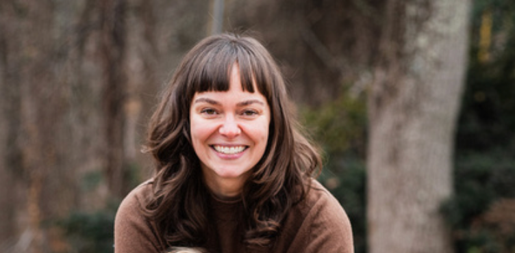 A woman with brown hair and a smile, wearing a brown sweater, outdoors in front of trees.