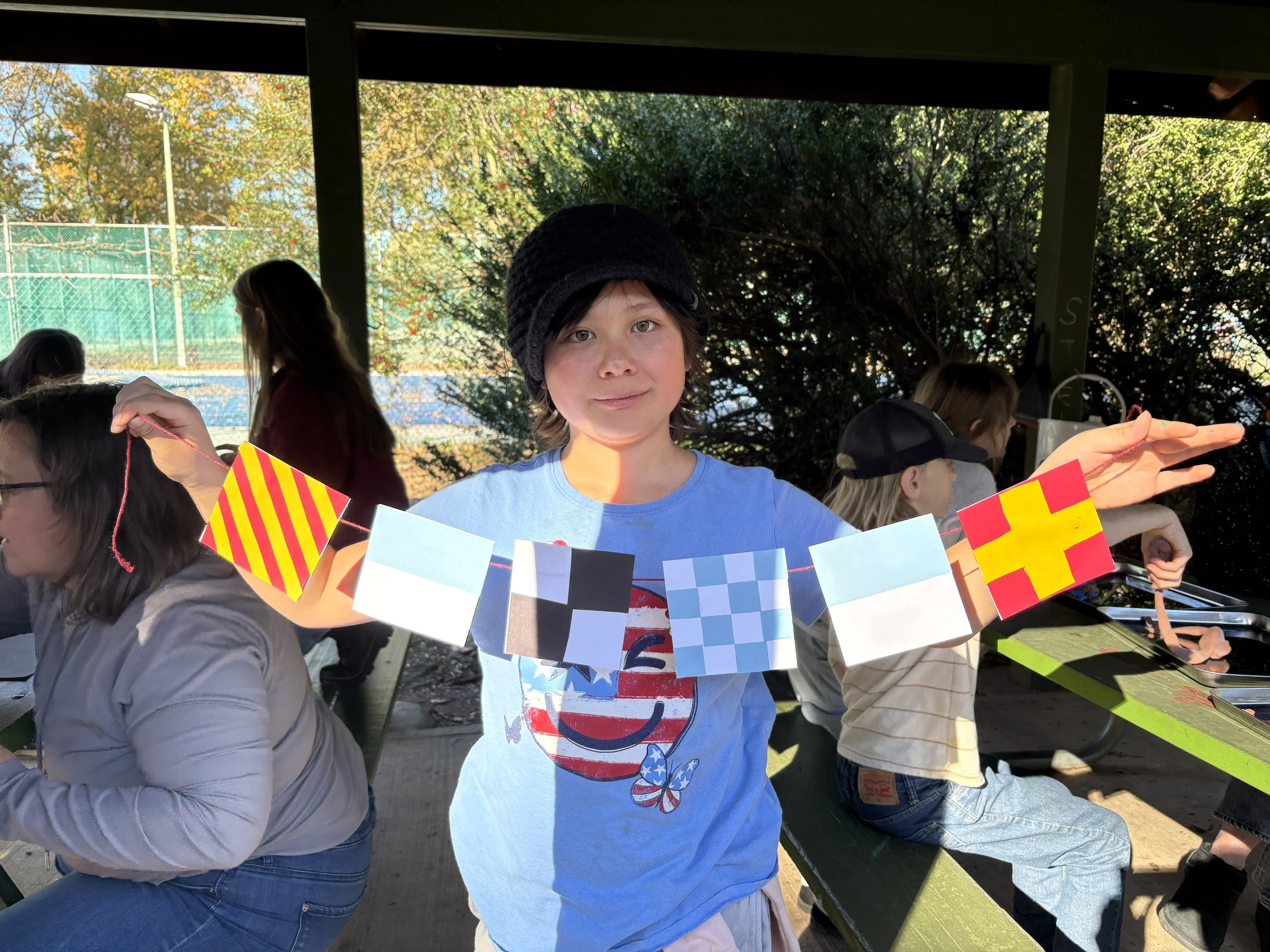 Child holding a string with colorful flags at an outdoor gathering, with other children and people in the background near tennis courts and trees.