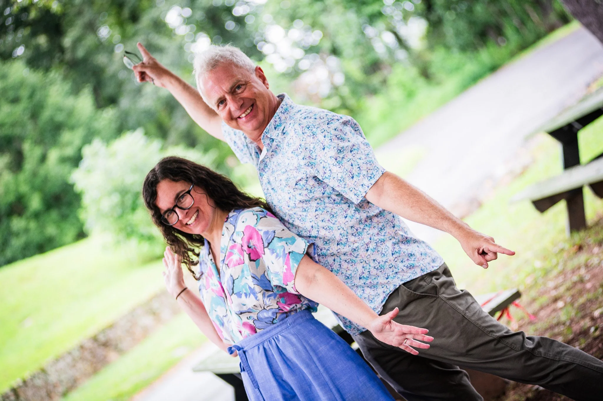 A man and woman smiling and pointing outdoors in a park with green trees in the background.