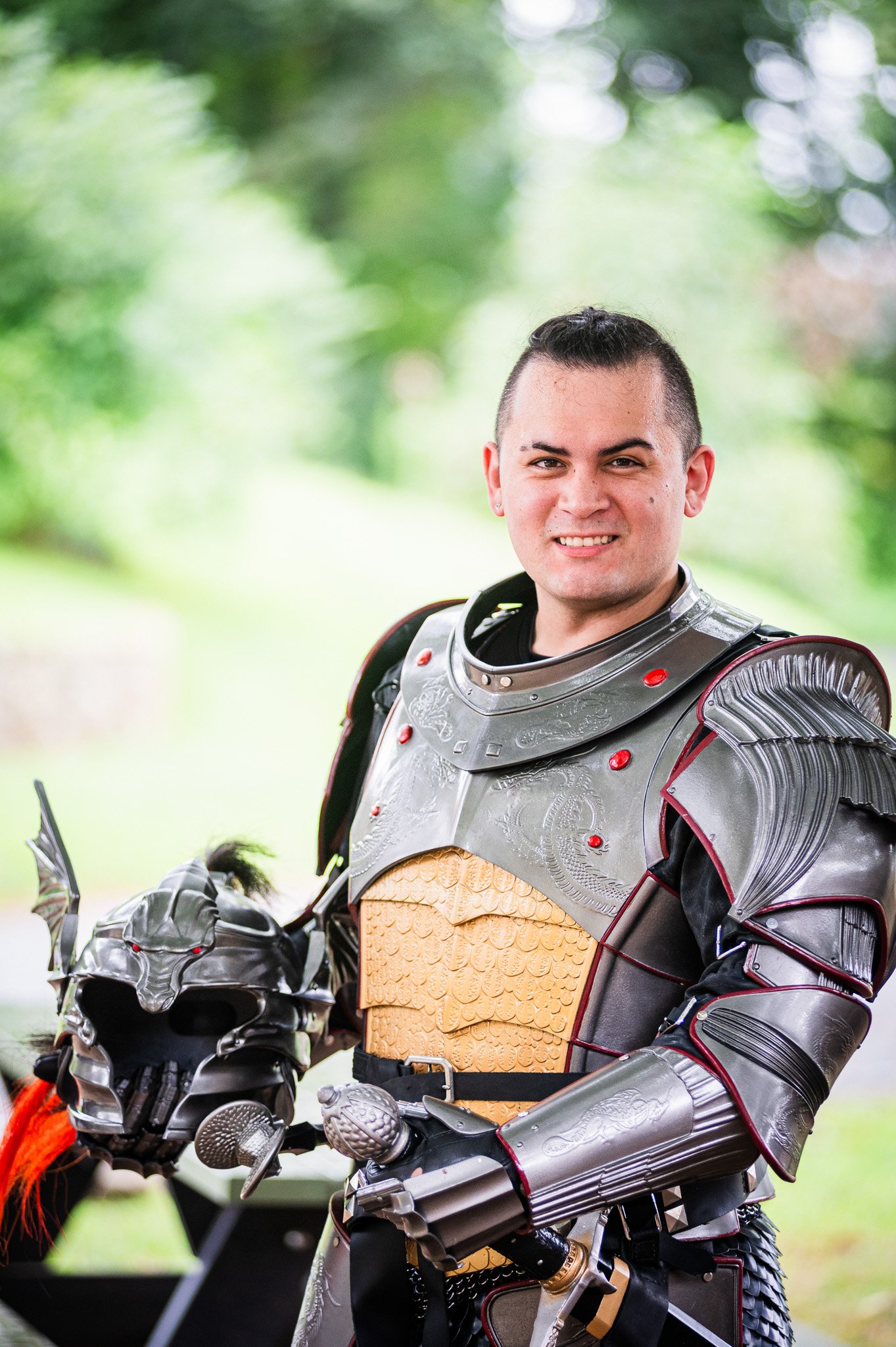 A man in cosplay warrior armor holding a decorative helmet, standing outdoors in front of blurred green trees.